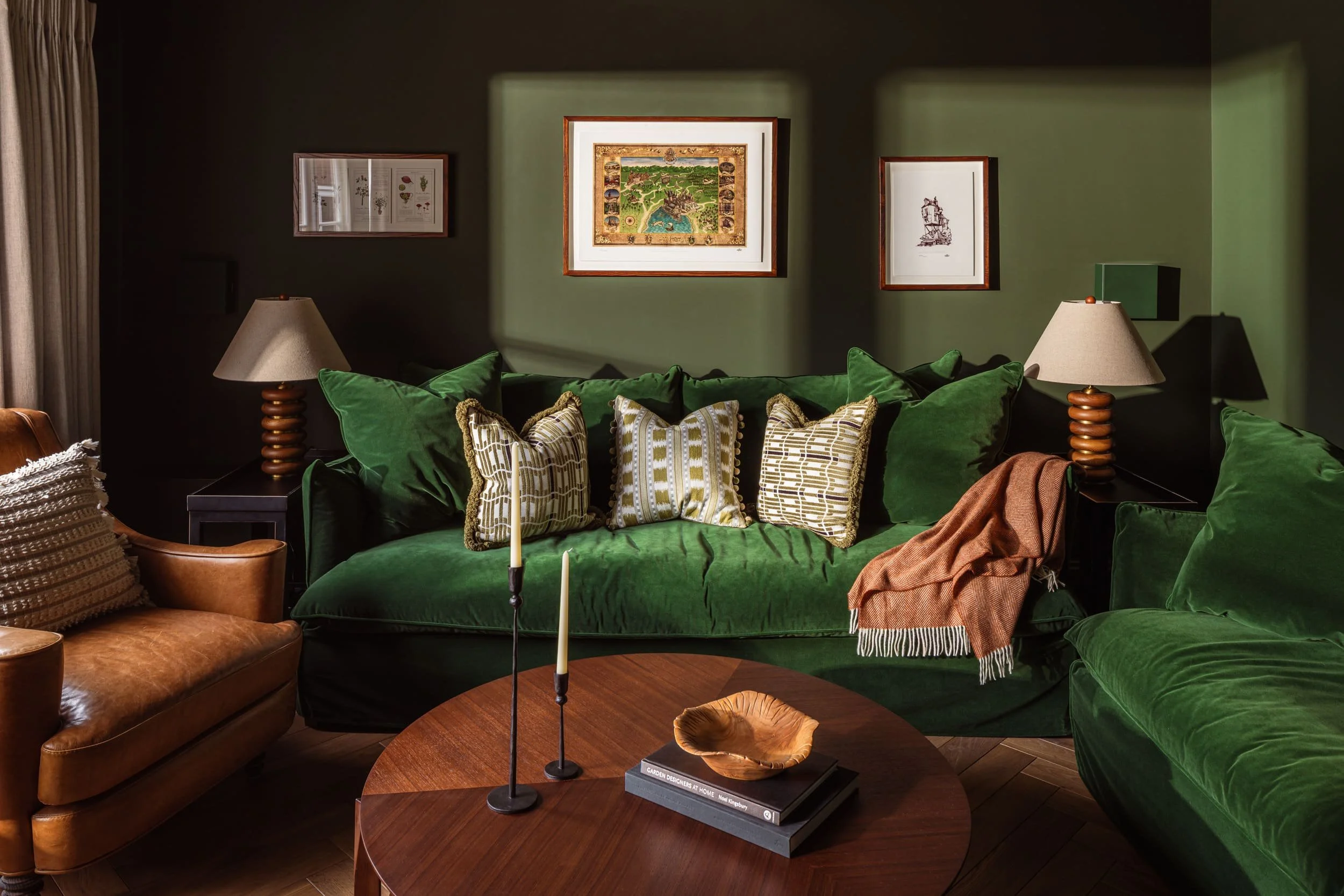 Living room with green velvet sofa, patterned throw pillows, wooden side tables with lamps, framed art on green walls, and a round wooden coffee table with decorative items.
