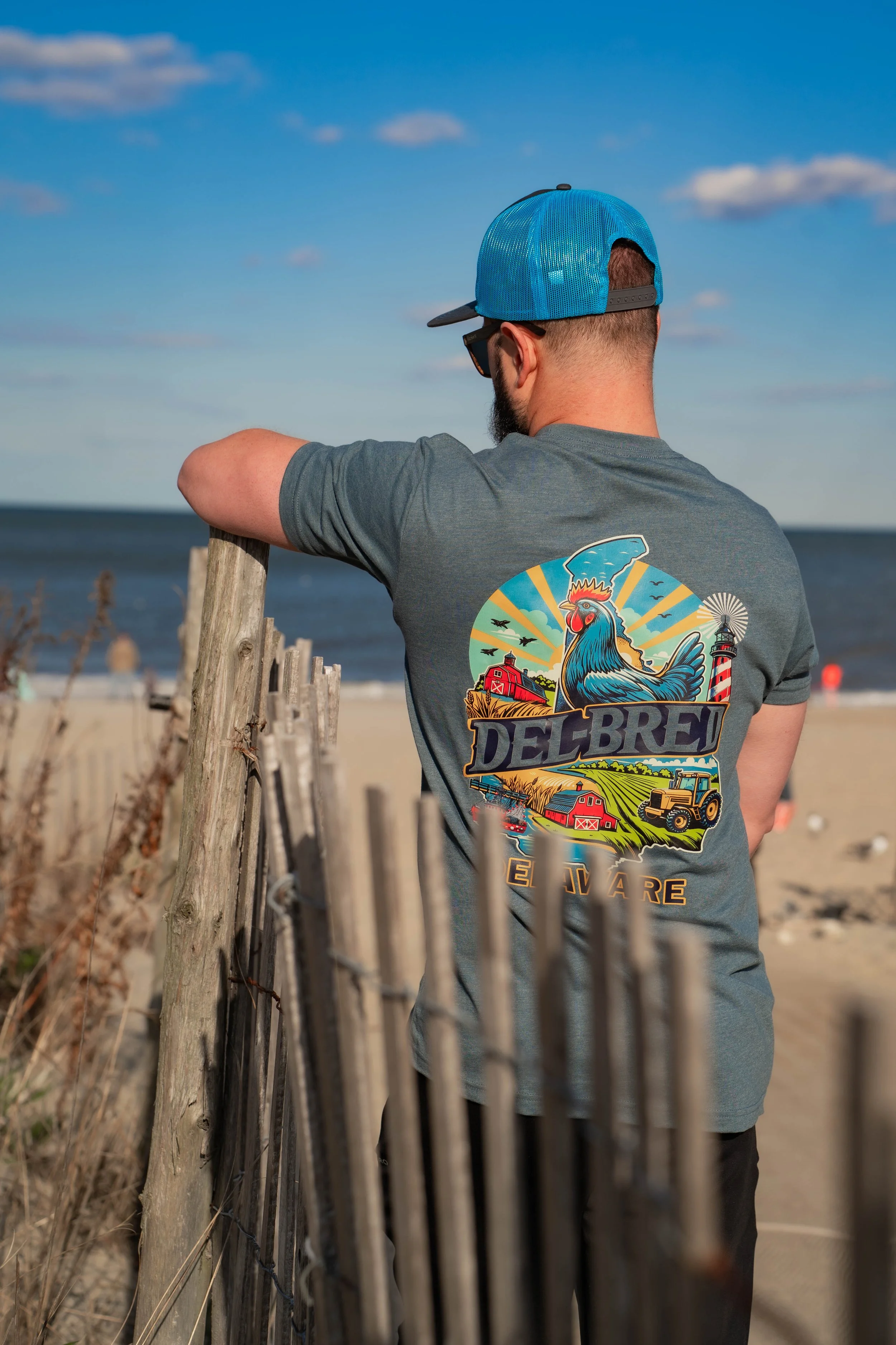 Man wearing a blue cap and aviator sunglasses standing on a beach, leaning on a wooden fence, looking at the ocean, wearing a gray t-shirt with a colorful graphic of a rooster, farm, and lighthouse.