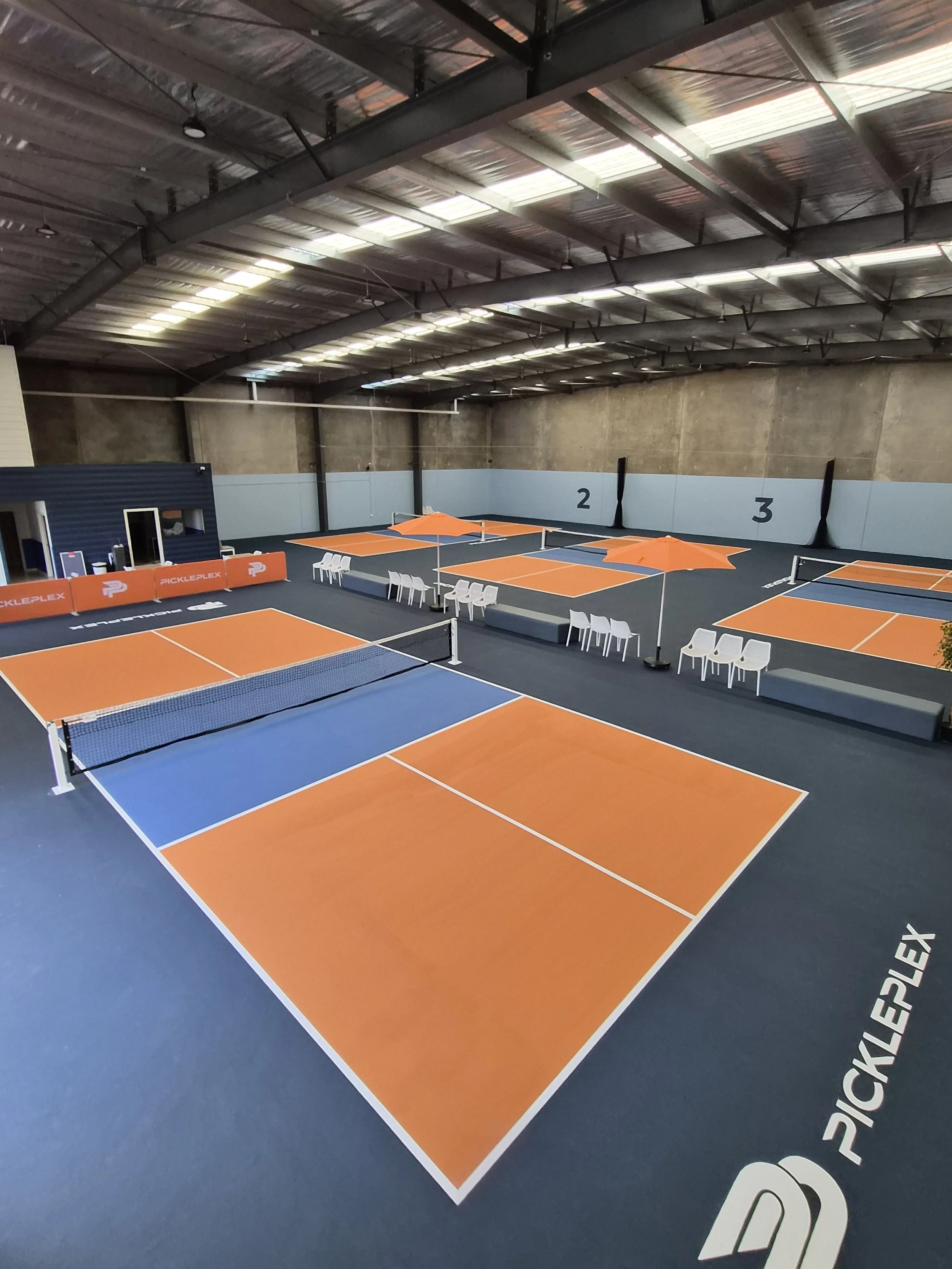 Indoor pickleball courts with orange and blue flooring, seating areas, and overhead lighting.