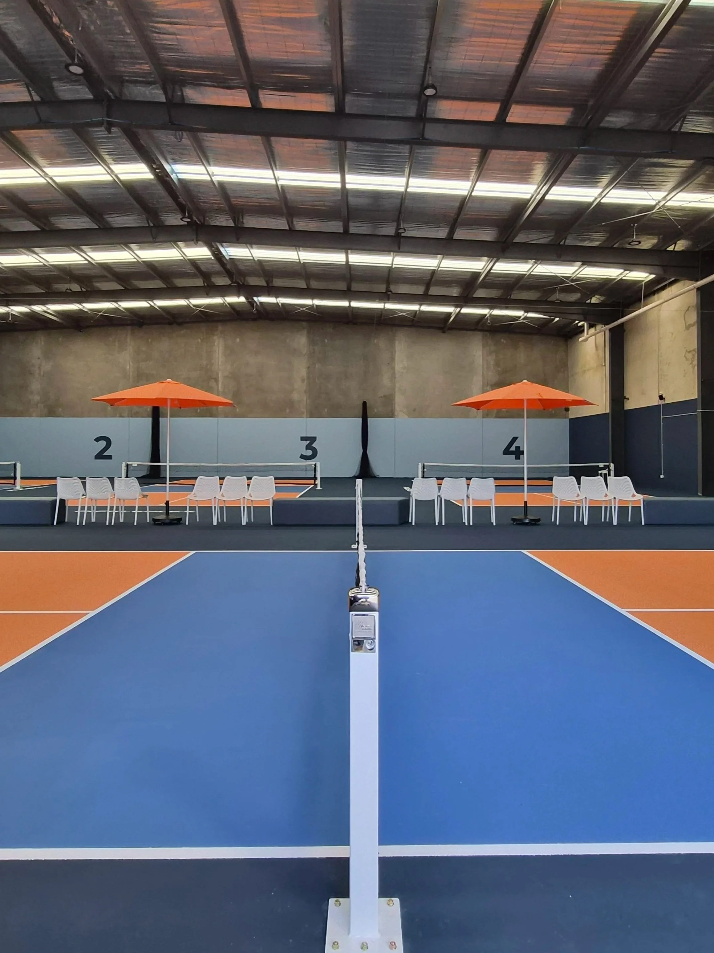 Indoor pickleball court with a net, blue and orange surface, spectator chairs, two orange umbrellas, and numbered dividing walls.