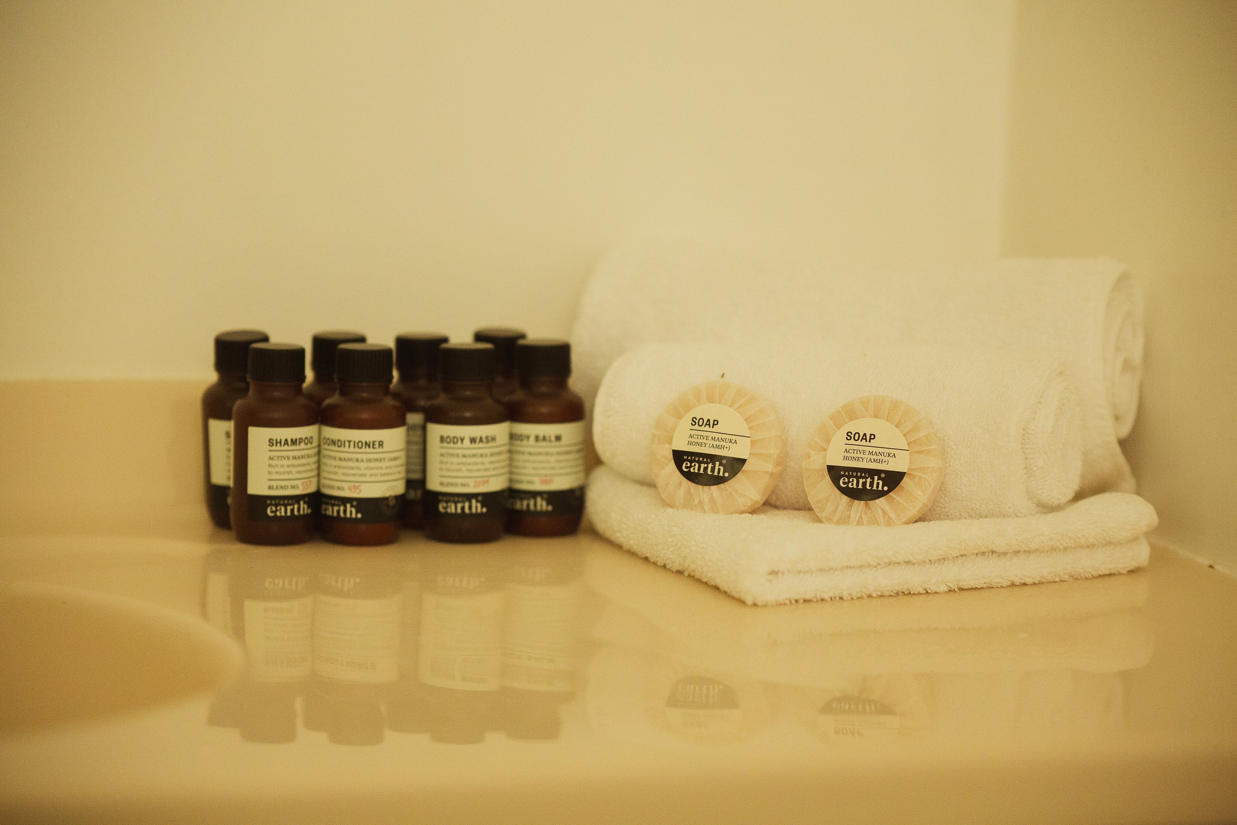 Spa or bathroom toiletries including bottles of shampoo, conditioner, body wash, and body balm, along with two circular soap bars labeled 'Active Manuka Honey' and a rolled white towel, all arranged on a surface with a mirror reflecting the items.