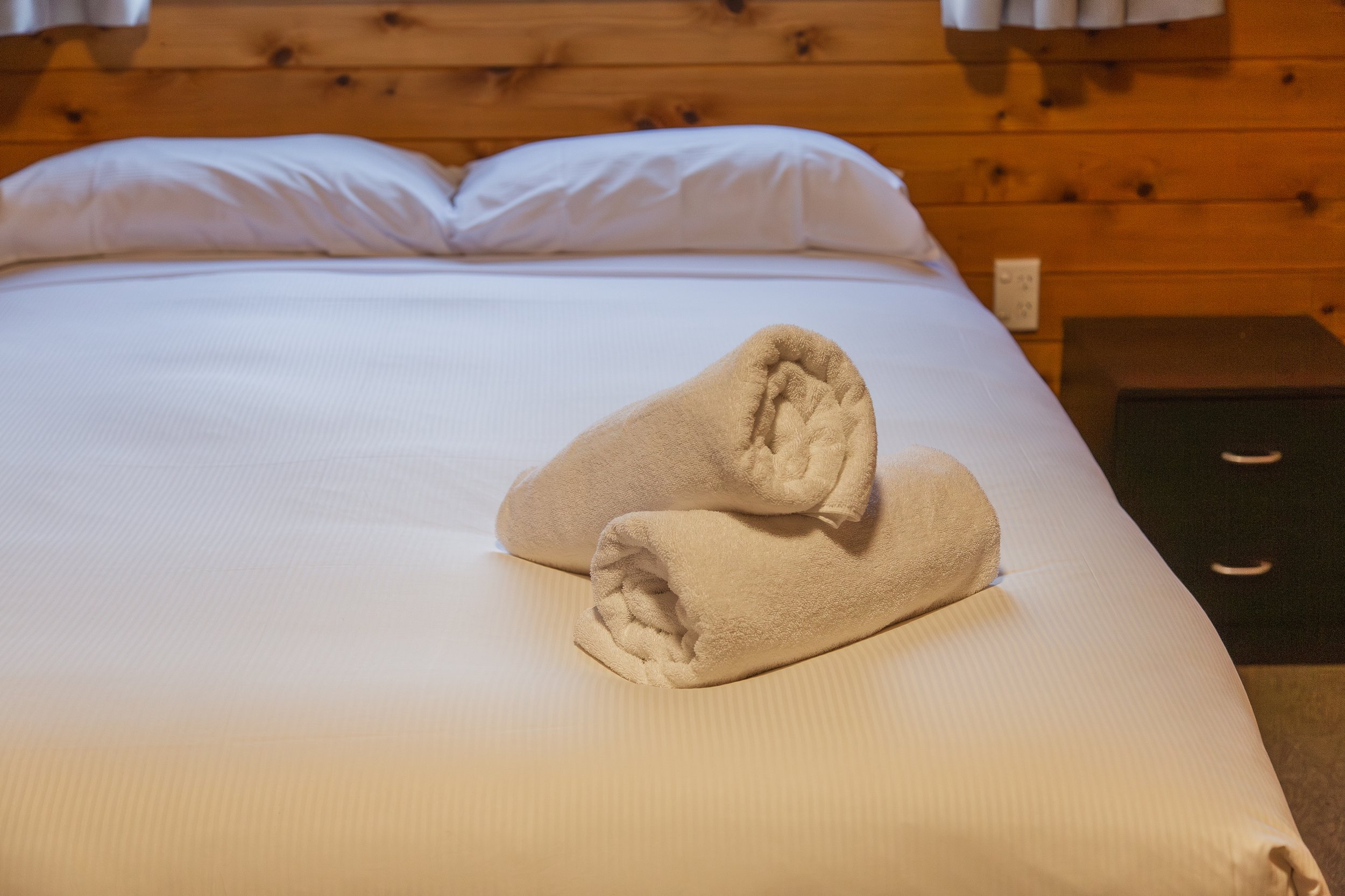 A neatly made bed with white linens and two rolled white towels on top, in a room with wood-paneled walls and a black bedside table.