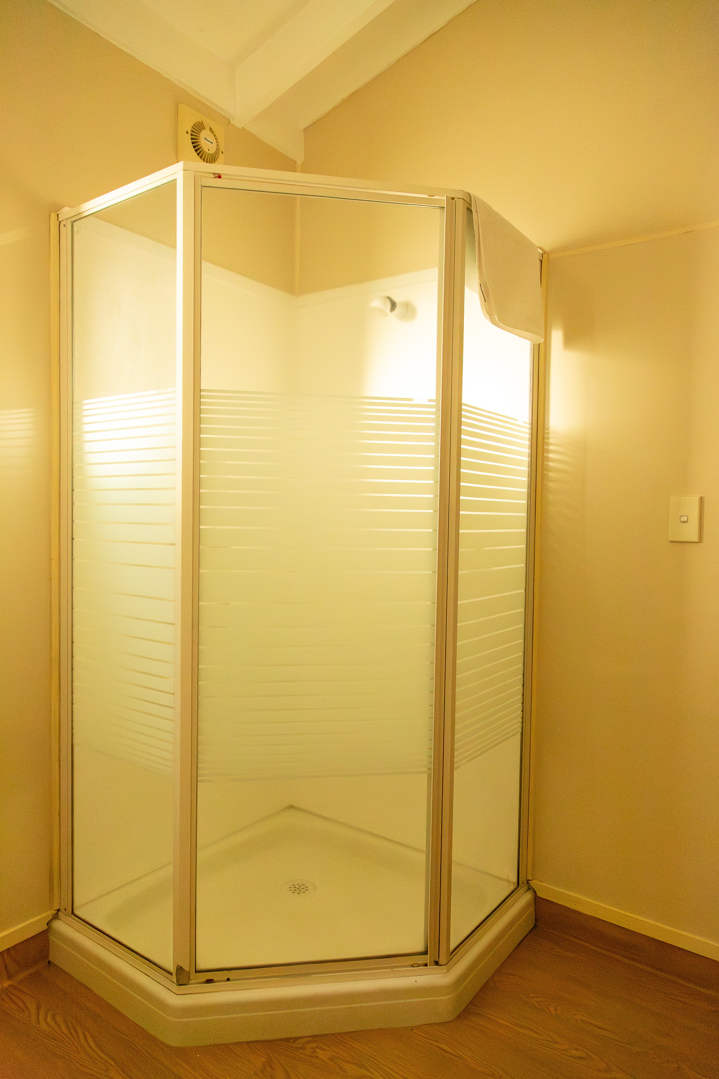 A corner shower stall with frosted glass panels and a beige frame, located in a room with wooden flooring and beige walls.