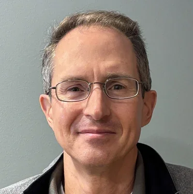 A middle-aged man with glasses and short gray hair, smiling slightly, against a plain gray background.