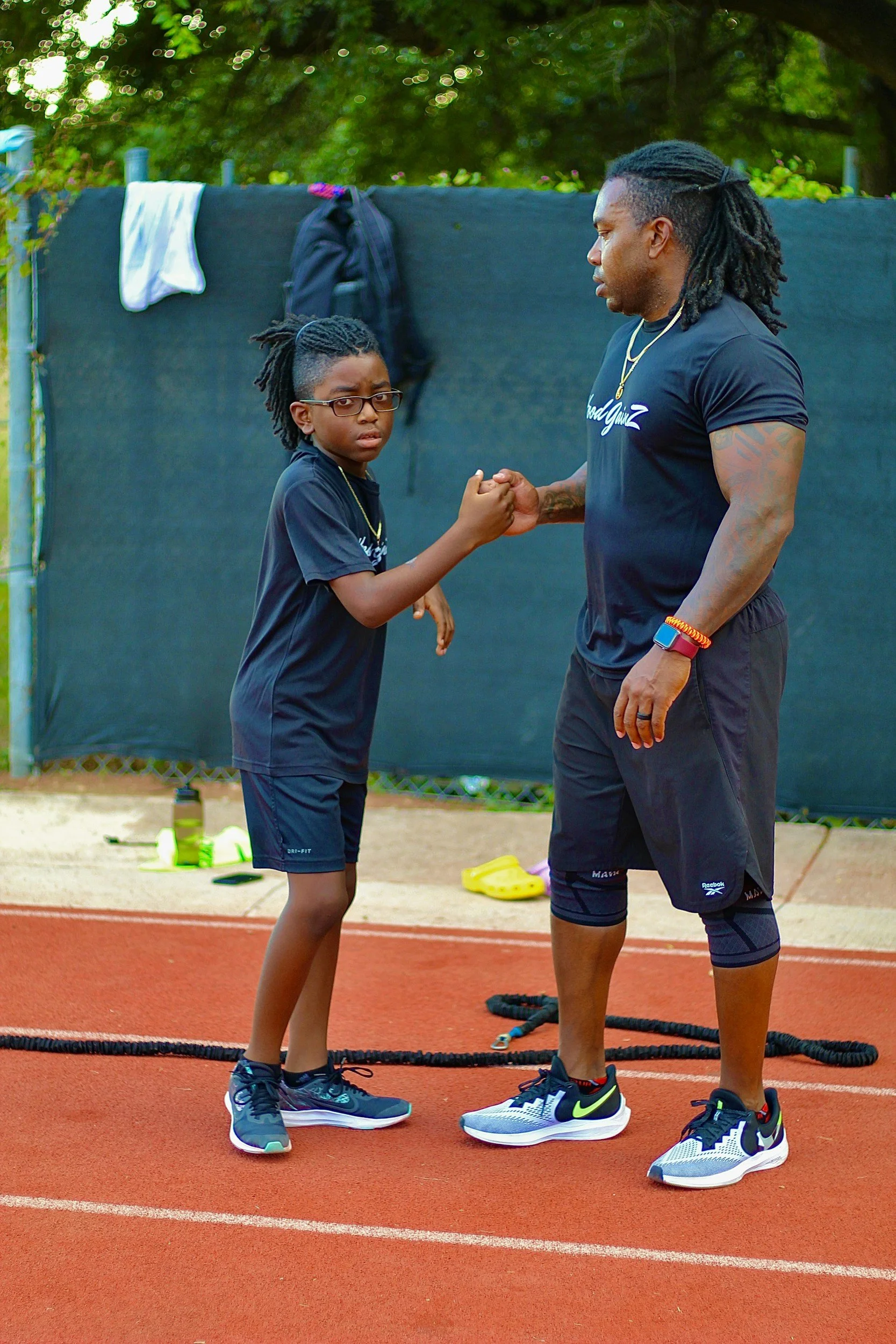 A man and a girl standing on a running track, holding hands with interlocked pinky fingers, outdoors near a black fence and trees. The girl is wearing glasses, a black t-shirt, black shorts, and running shoes. The man is dressed in athletic attire, with a black shirt, black shorts, and sneakers. They appear to be engaging in a handshake or a similar gesture.