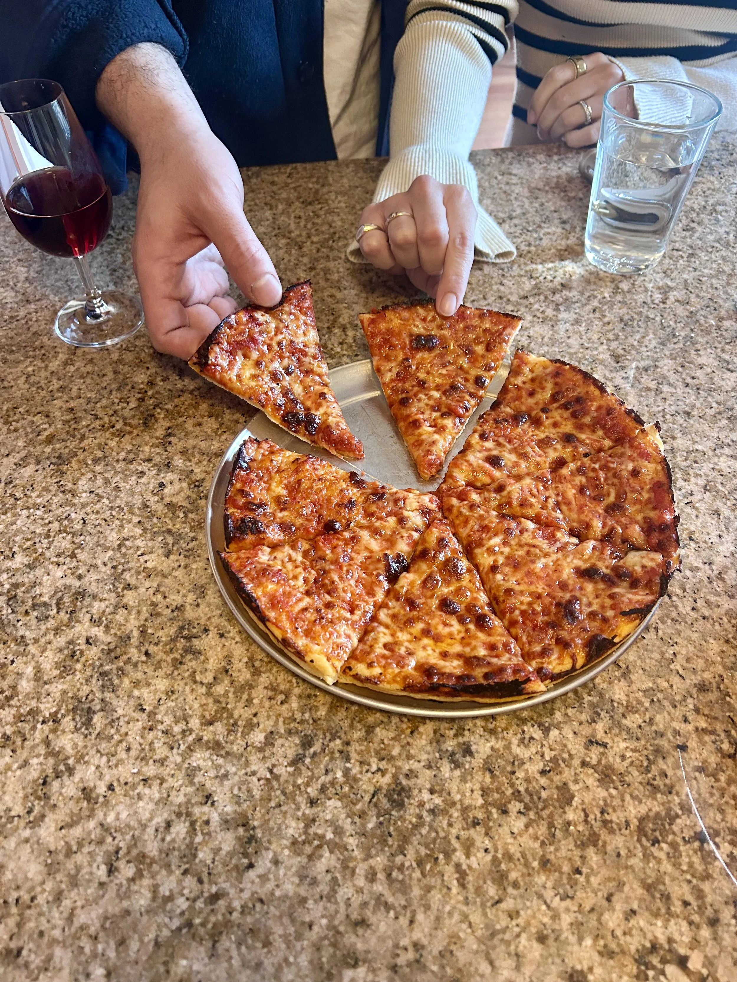 Two people at a table pointing at slices of cheese pizza on a plate. There is a glass of red wine and a glass of water on the table.