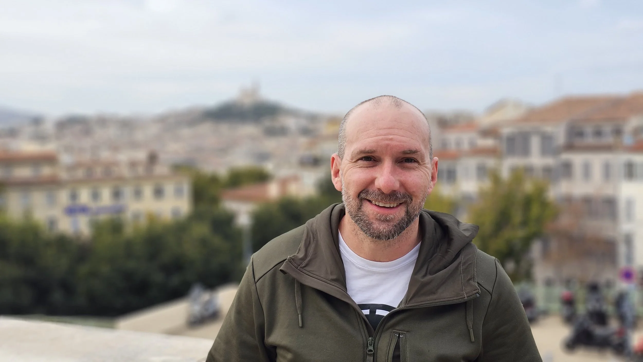 Smiling man with a beard and bald head wearing a green jacket, posing outdoors with a cityscape background featuring buildings and a hill.