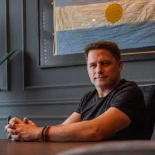 A man with short hair wearing a black T-shirt sitting at a table in a room with dark green walls and a window behind him.