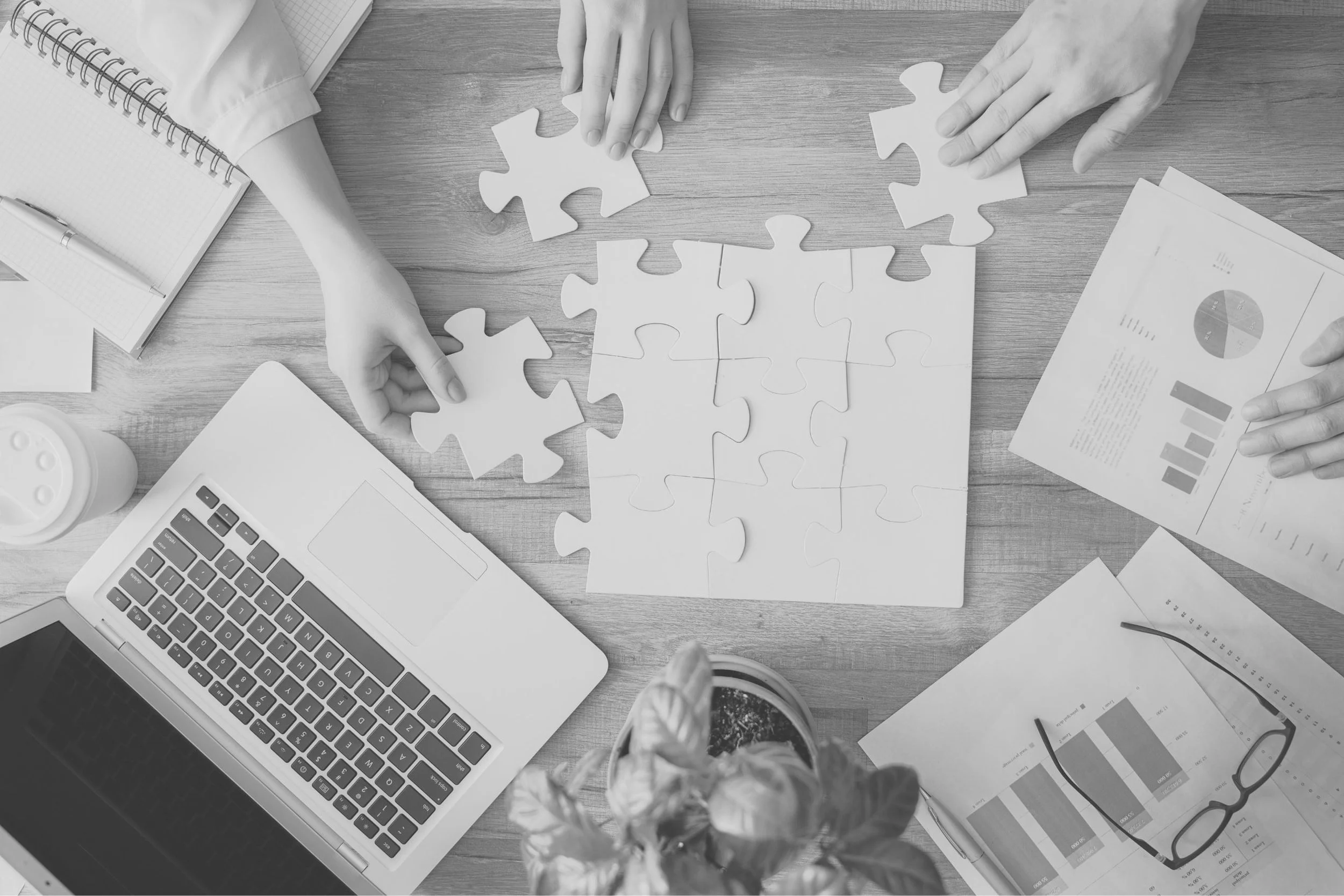 Drei Personen arbeiten an einem Puzzlespiel aus Papier auf einem Schreibtisch mit Laptop, Notizbüchern, Diagrammen, einer Brille, einem Pflanzenstück und einem Medikamentenflaschen.