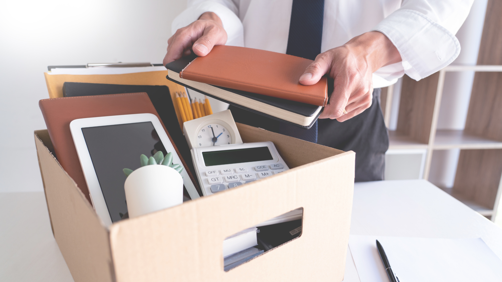 Person packing a cardboard box with office items such as notebooks, a tablet, a plant, a clock, a calculator, and pencils.