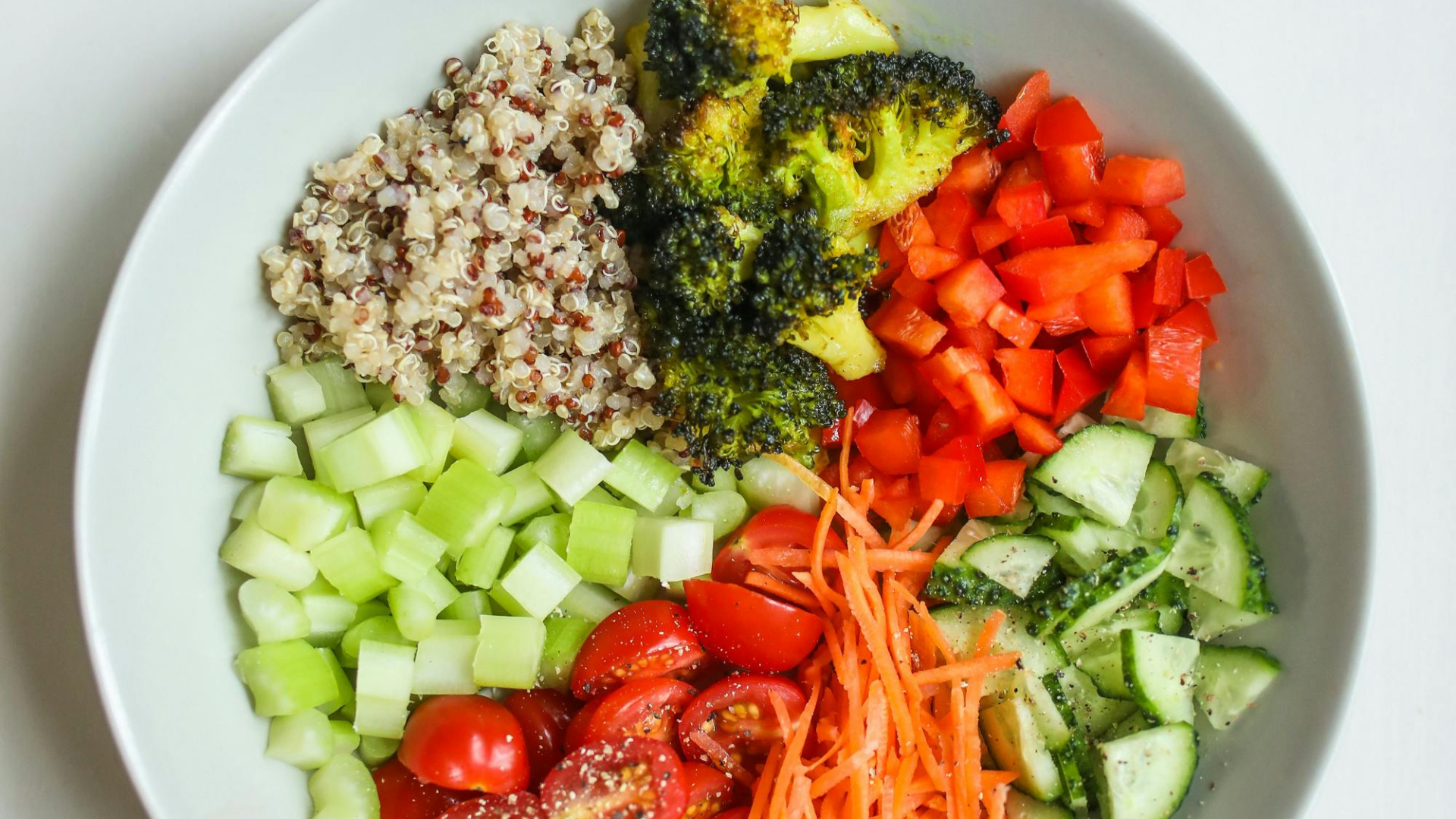 A white bowl filled with various chopped vegetables and grains, including cherry tomatoes, cucumber, grated carrot, red bell pepper, broccoli, cooked quinoa, and diced celery.