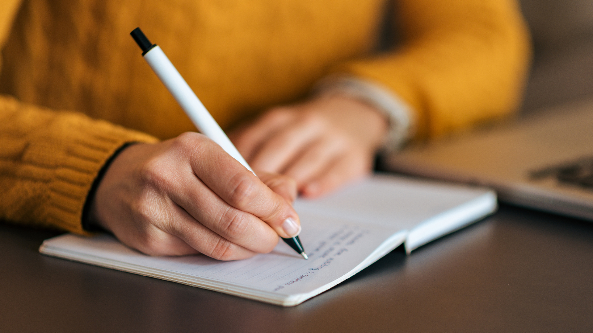 Person wearing a yellow sweater writing in a notebook with a pen, with a blurred laptop in the background.