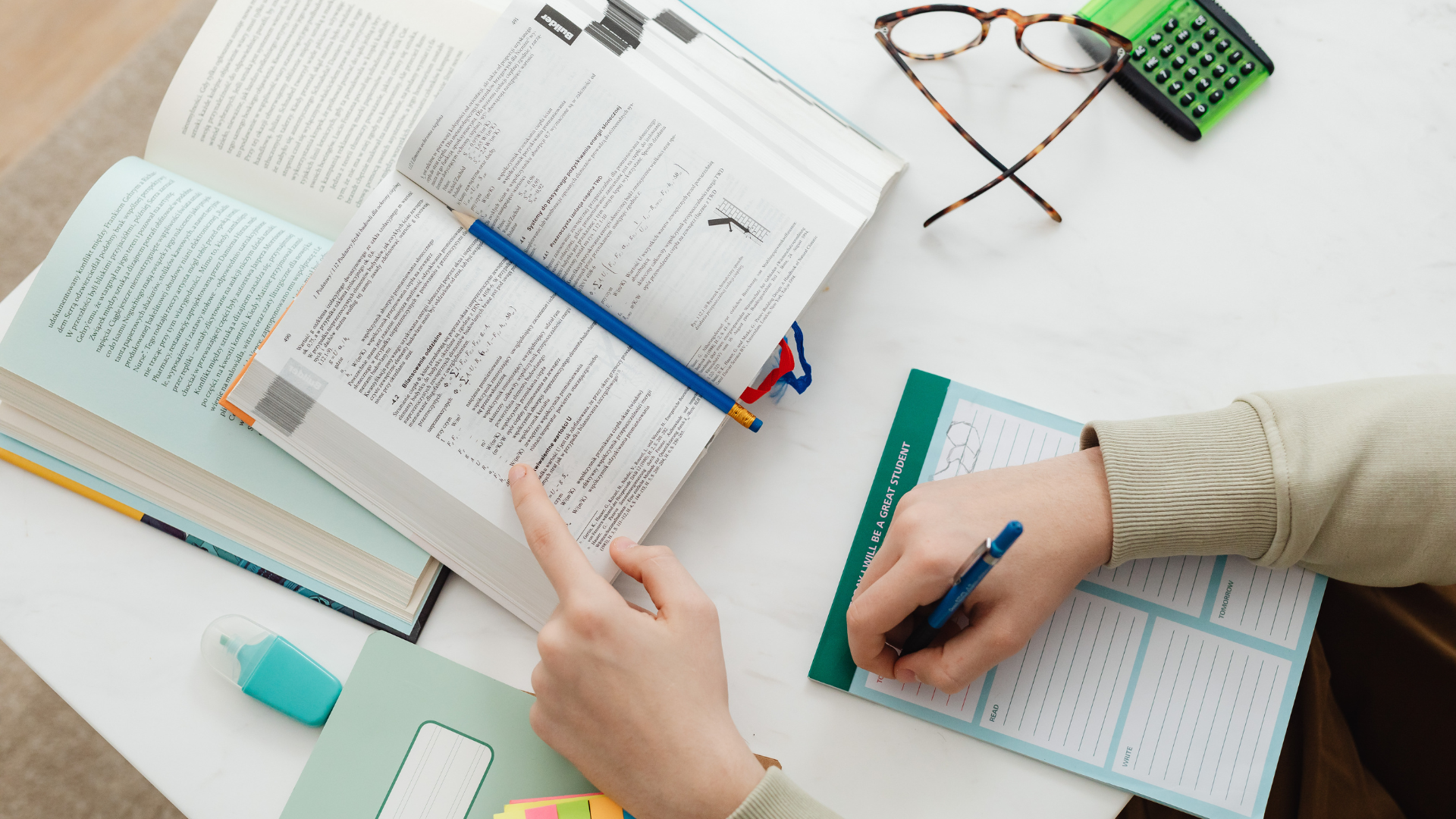 Person studying with open textbooks, a blue pen, a flash drive, colorful sticky notes, and a note sheet on a white desk, with glasses and a calculator nearby.