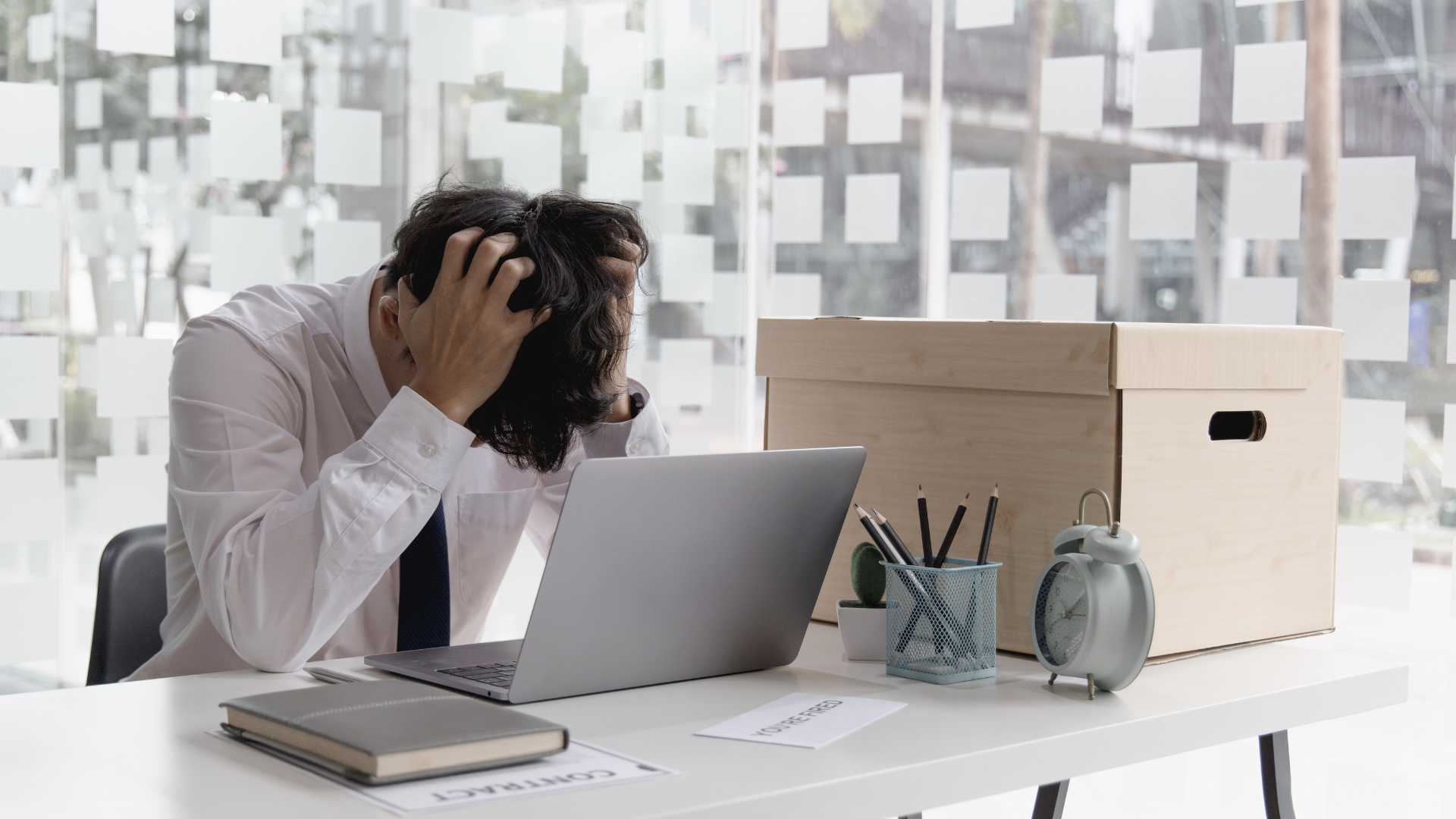 Frustrated person holding head in office with a laptop, notebook, container with pens, a clock, and a wooden box on the desk.