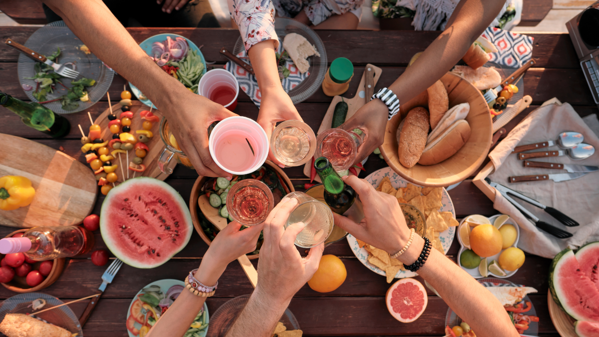 A group of people enjoying a barbecue or picnic, clinking glasses with drinks over a wooden table filled with various foods including watermelon, bread, olives, vegetables, chips, and fruits.