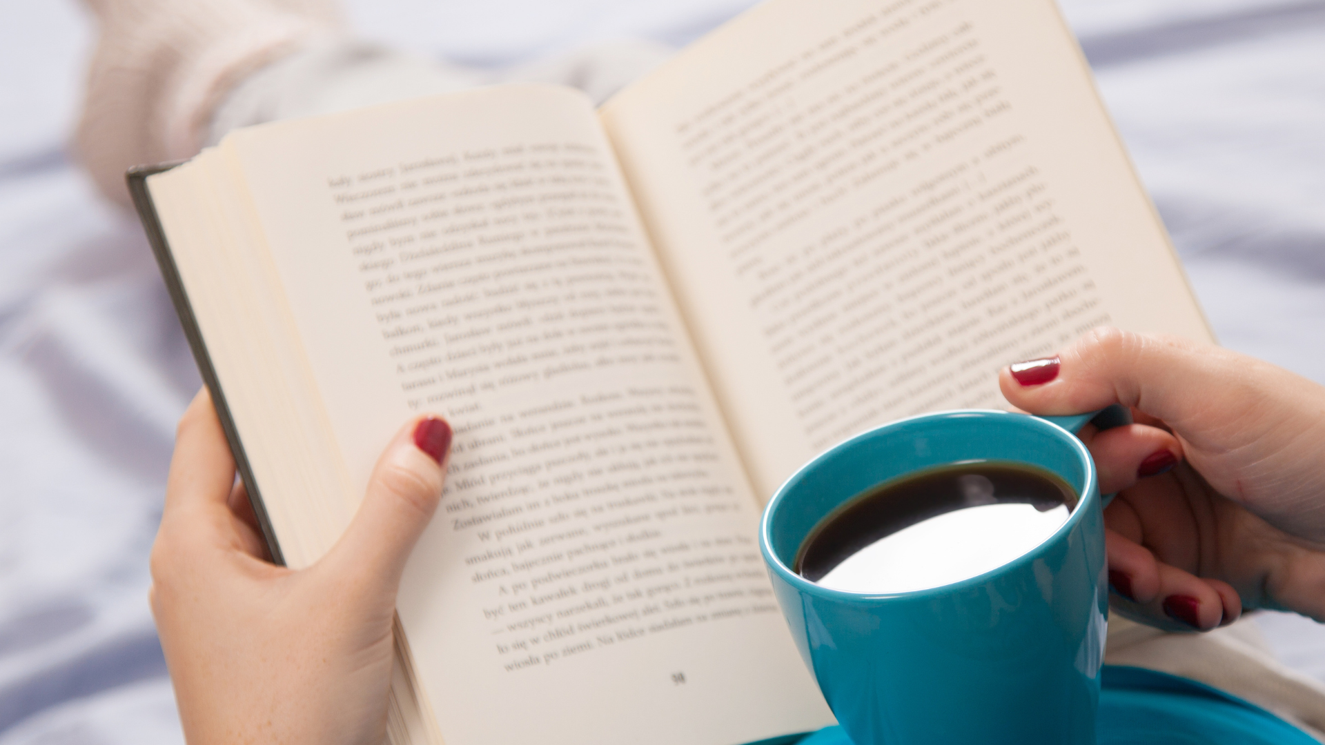 Person holding an open book in one hand and a blue cup of coffee in the other, with a blurred background.