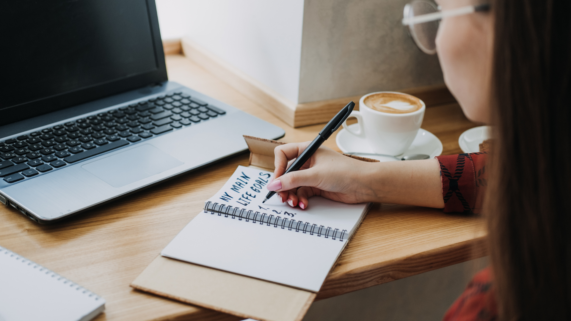 A person with glasses writes in a notebook on a wooden desk, with a laptop, a cup of coffee, and a biscuit nearby.