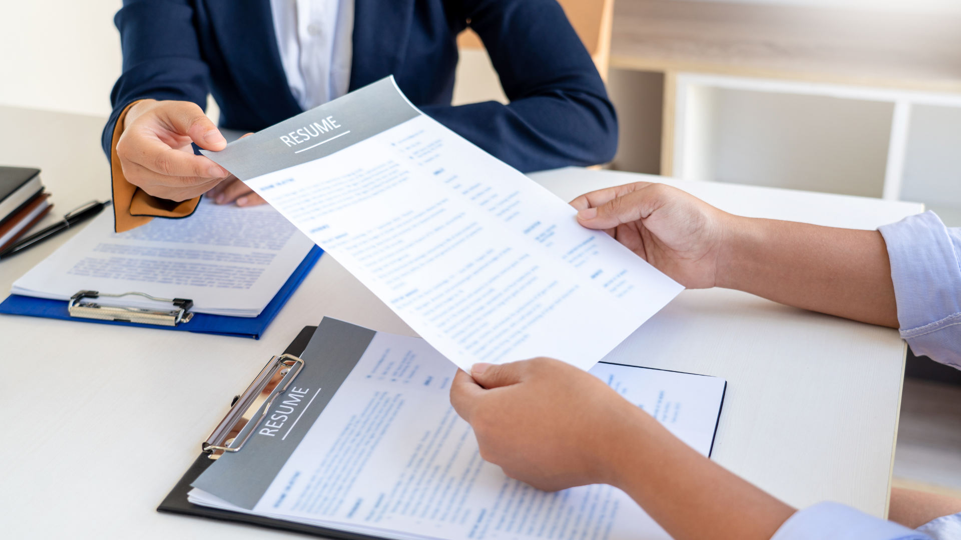 Person in business attire handing a resume to another person at a desk in an office.