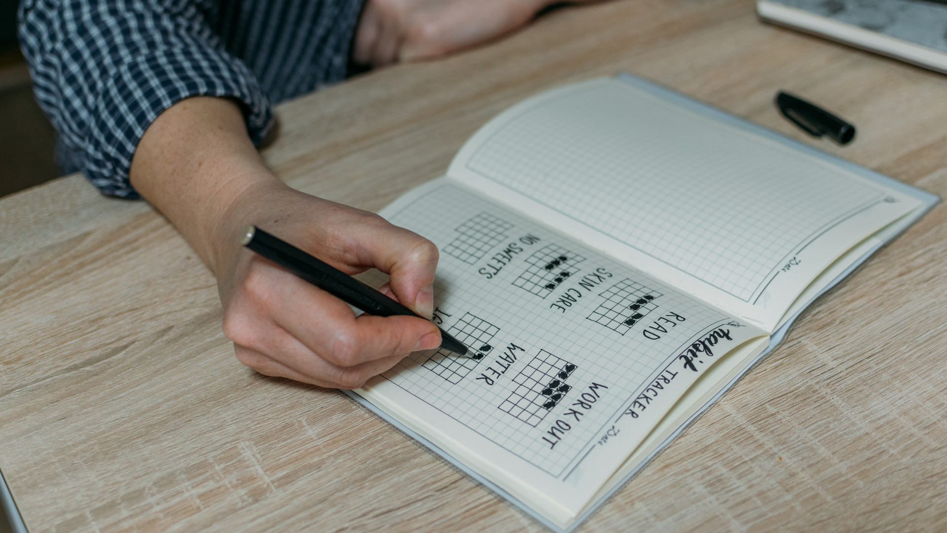 Hand writing on a grid notebook with music chord diagrams and song labels, on a wooden table.