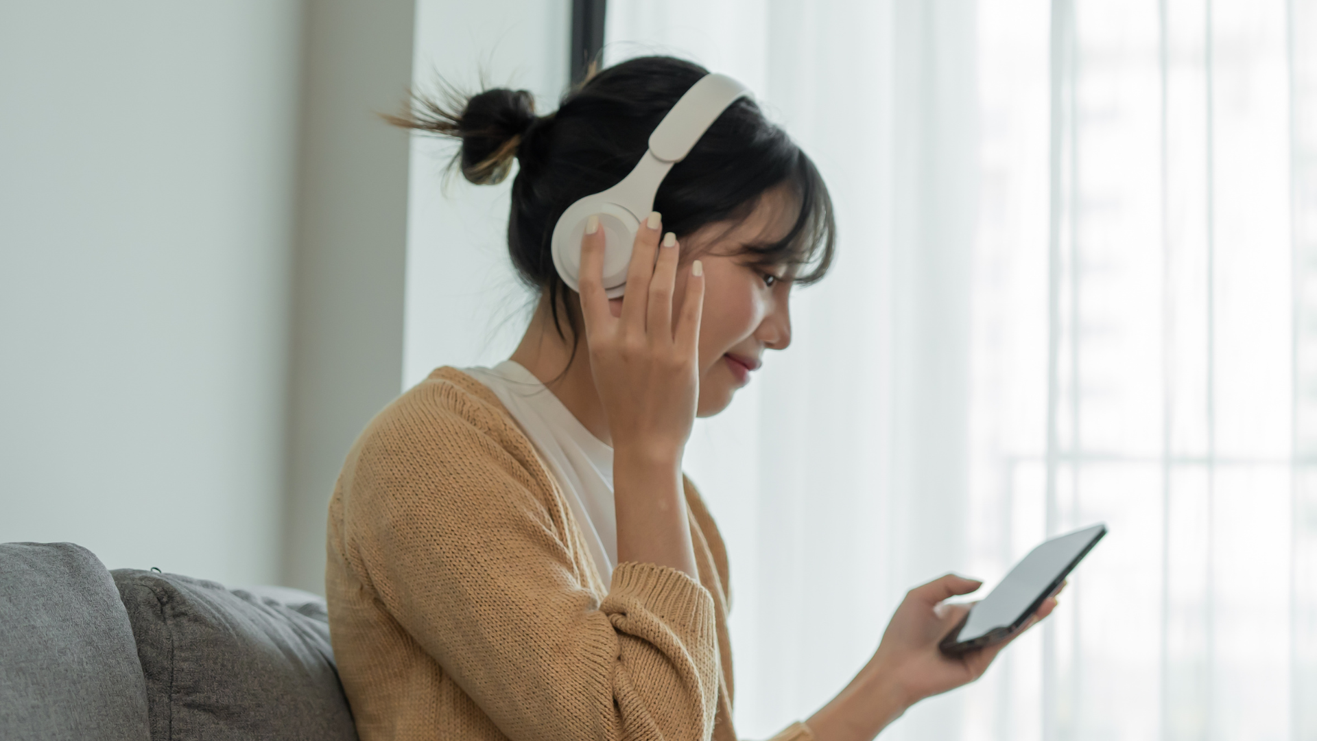 Young woman wearing white headphones, sitting on gray couch, looking at her phone, in a well-lit room with sheer white curtains.