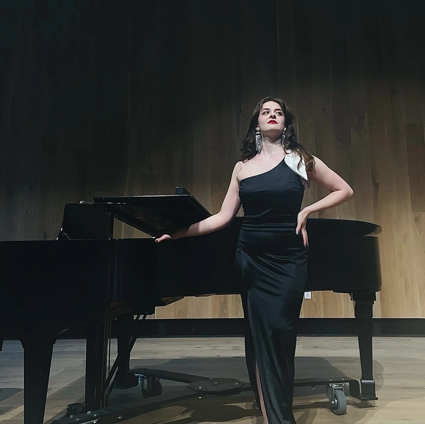 A woman in an elegant black gown with a white accent over one shoulder, standing next to a grand piano on a wooden stage with a dark wood wall background.