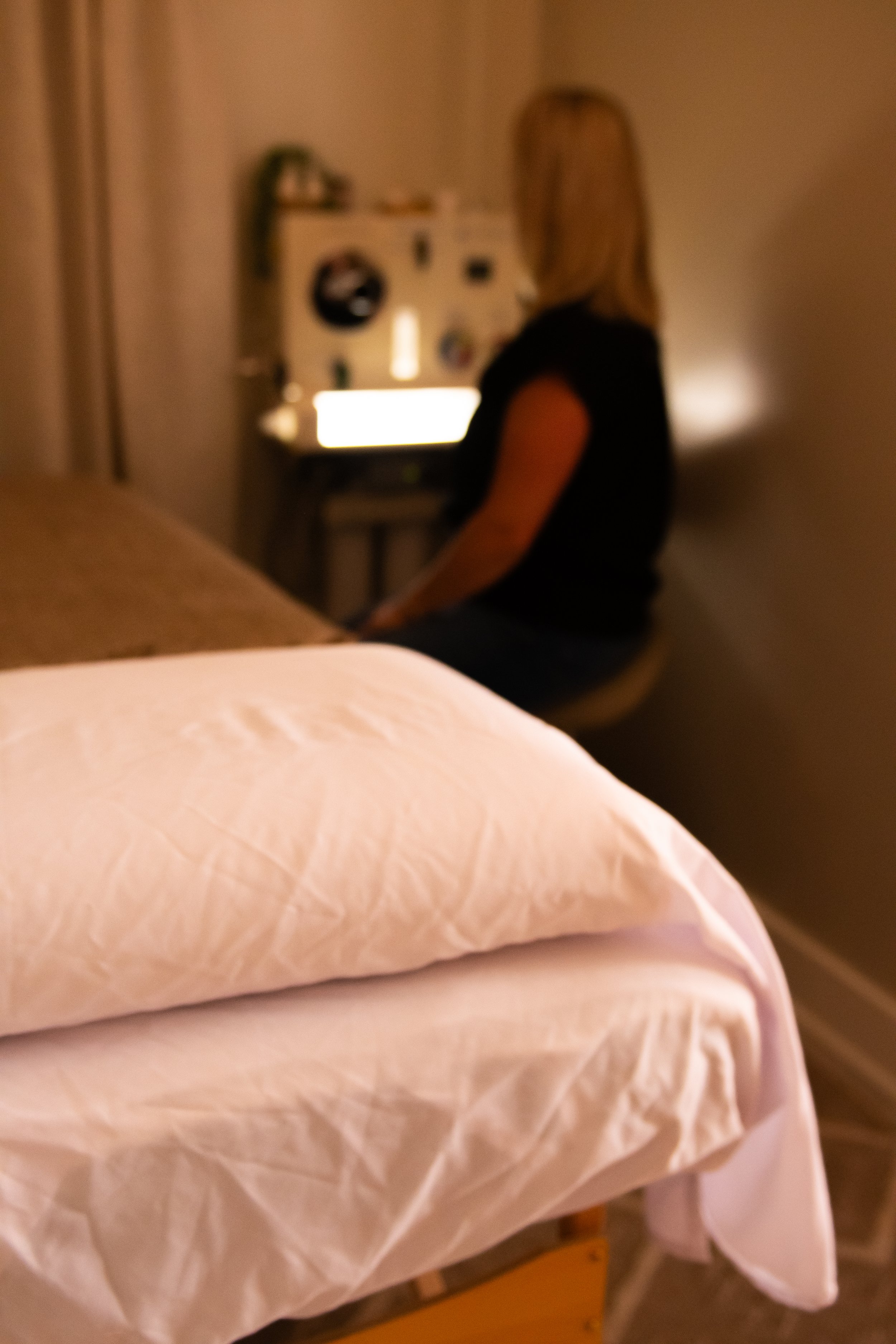 A slightly out-of-focus woman with blonde hair sitting at a desk or table, facing to the right, in a dimly lit room, with a bed and pillow in the foreground.