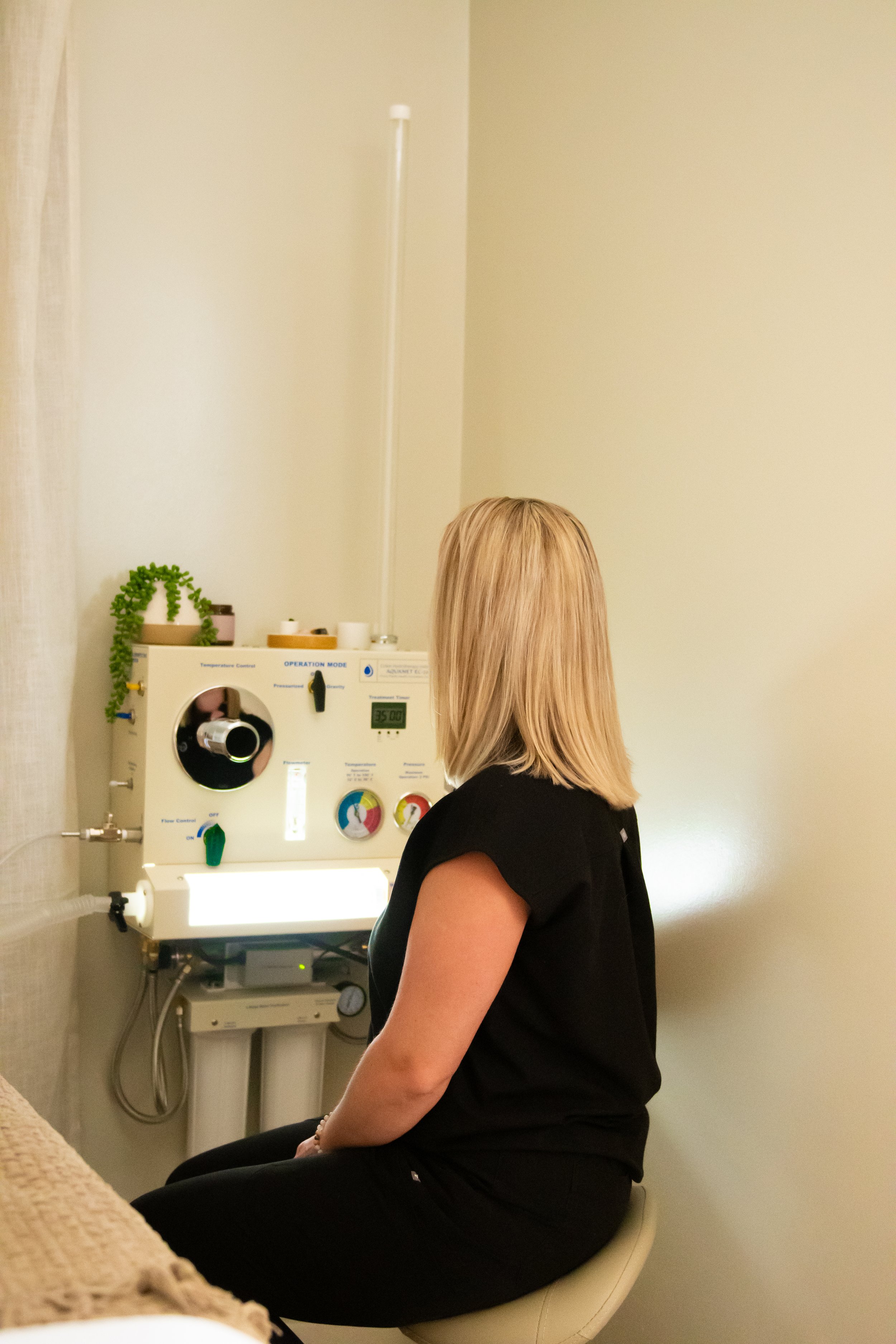 A woman sitting on a stool in a medical room, facing a medical device with various controls and displays, for respiratory or oxygen therapy.