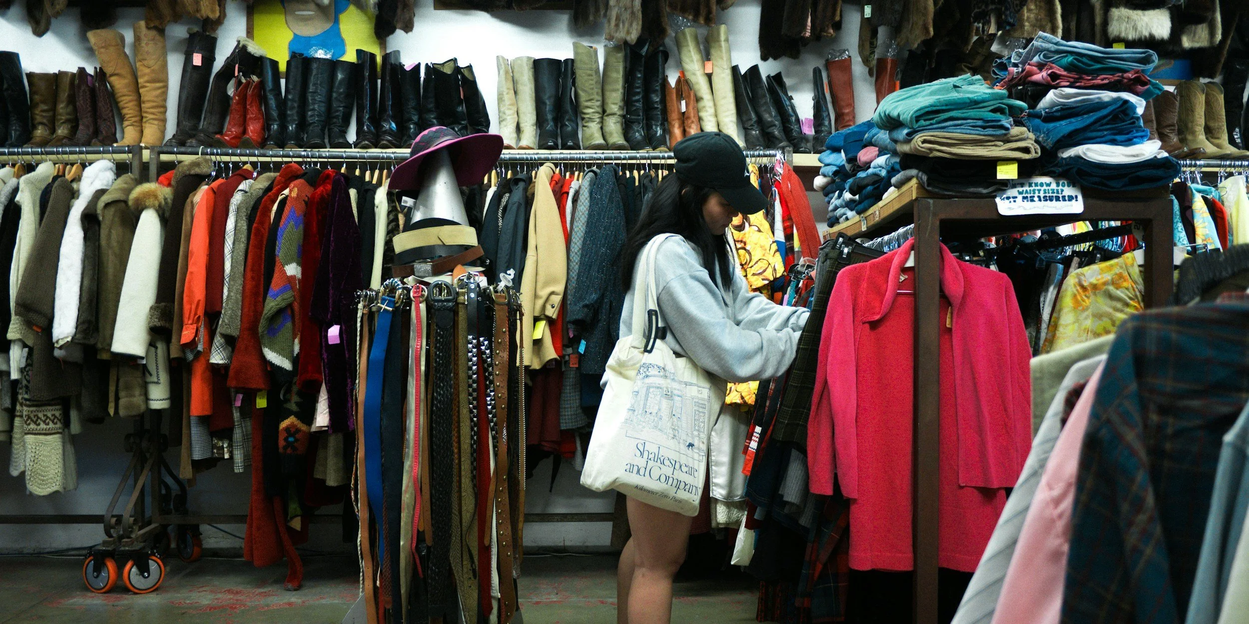 A woman shopping for clothes in a thrift store, surrounded by racks of jackets, shirts, and boots on the wall.