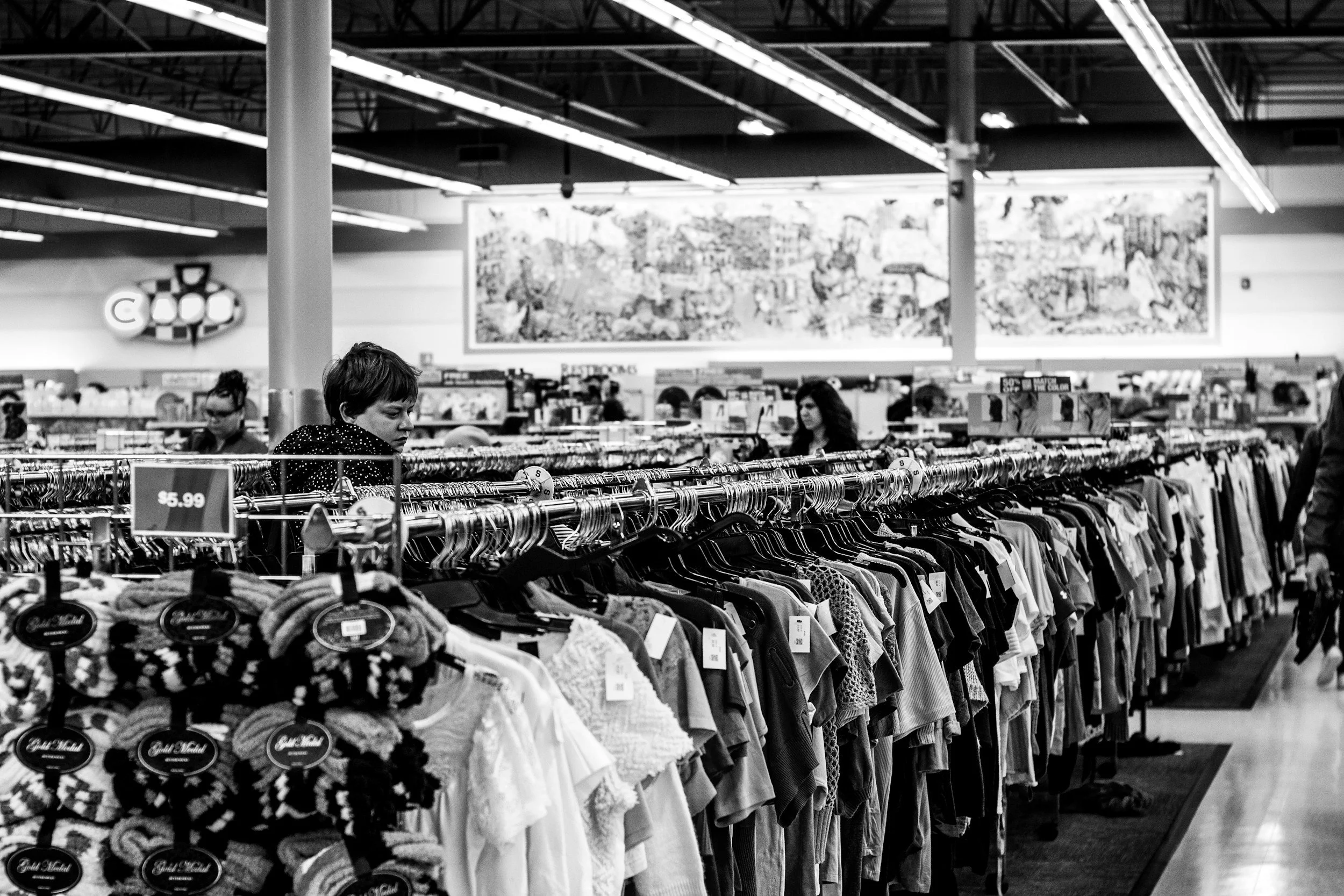 Black and white photo of a store aisle with clothing racks filled with various garments. Two women are shopping, one in the foreground and another further down the aisle. Bright ceiling lights, a large wall art piece, and signs indicating departments like 'Restrooms' and price tags are visible.