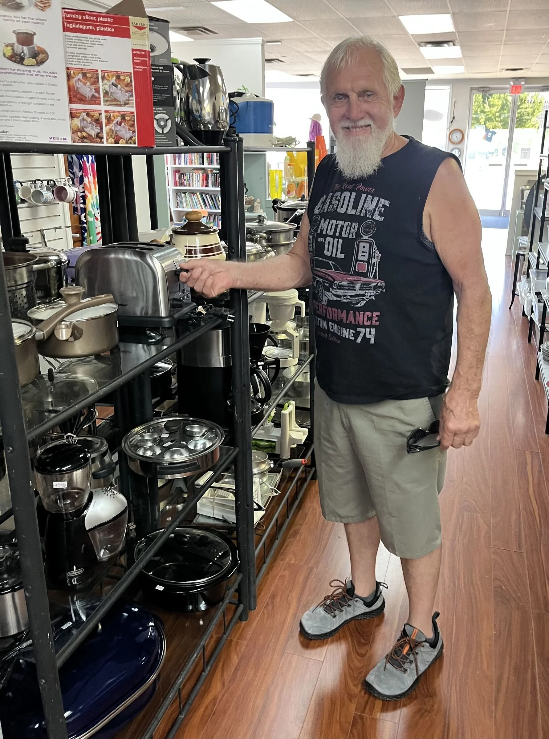 A man with a white beard, wearing a sleeveless black t-shirt with a car graphic and khaki shorts, stands in a store among kitchen appliances and cookware, holding a pair of sandals.
