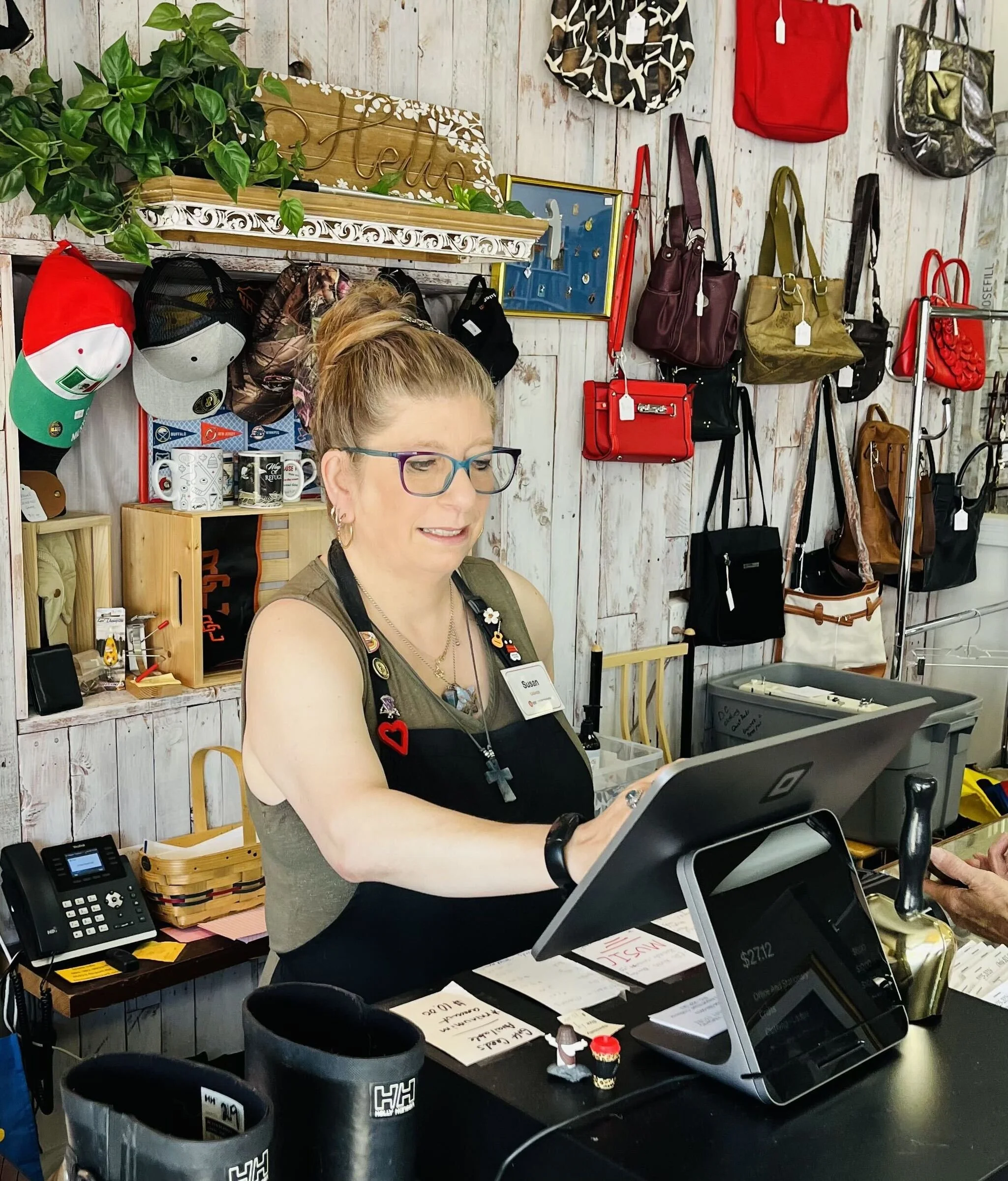 A woman with glasses and a name tag working at a checkout counter in a store, with various handbags and accessories hanging on the wall behind her.