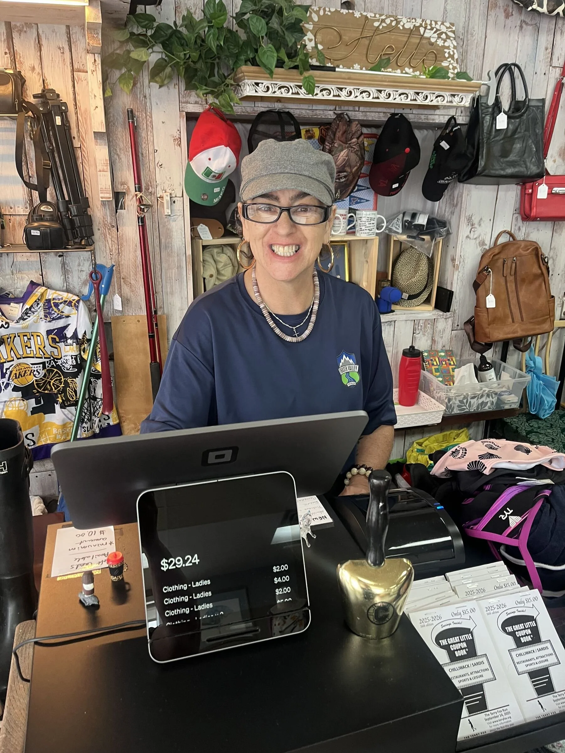 A woman smiling at a checkout counter in a store. She is wearing glasses and a light gray cap, and has earrings. The counter has a black cash register, brochures, and a gold-colored trophy. In the background, there are shelves with hats, hats with tags, a plant, and decorative items.