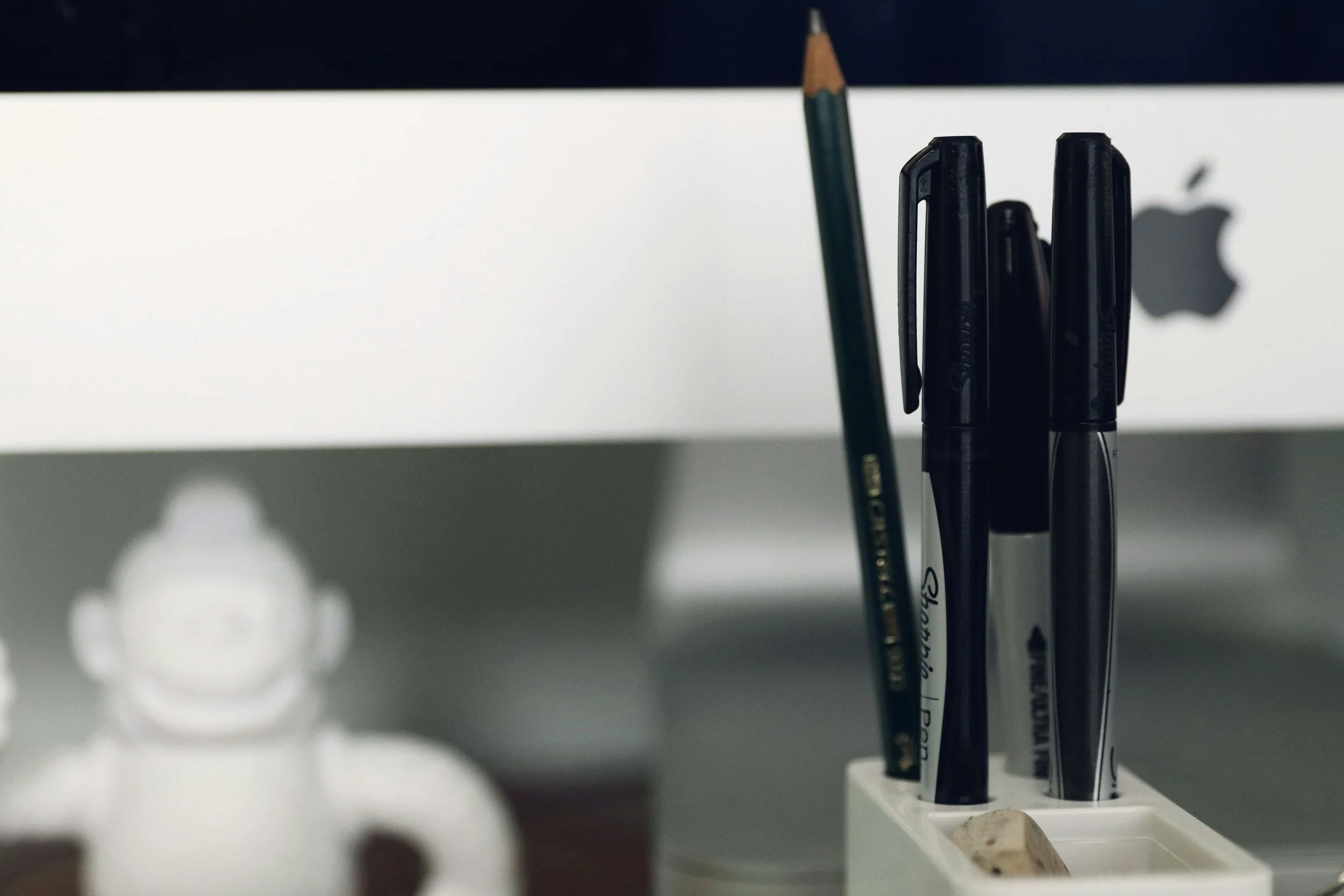 Lápiz y varios bolígrafos en un portalápices en un escritorio, con un fondo difuso de una pantalla de computadora.