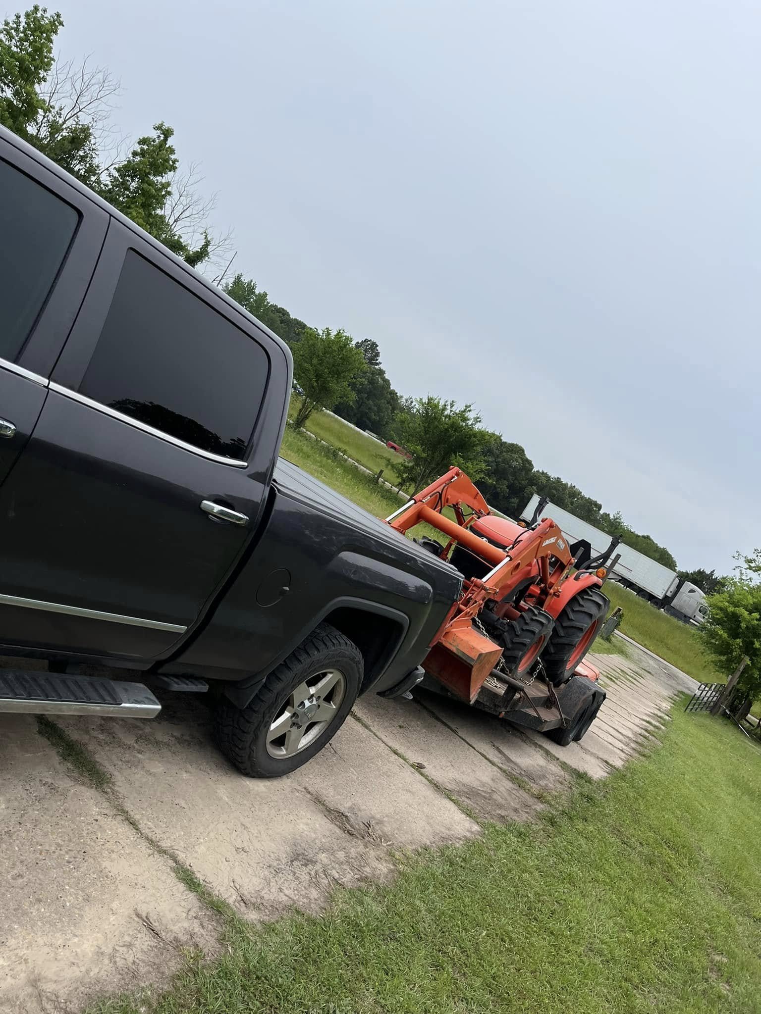 A black pickup truck transporting an orange tractor on a flatbed trailer parked on a concrete driveway in a rural area with green grass, trees, and a cloudy sky.