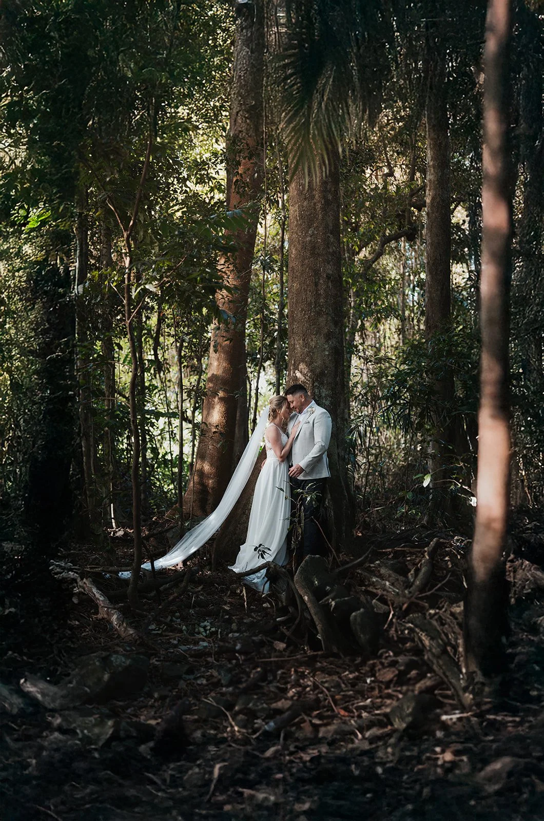 A bride and groom stand close together in a rainforest, gazing at each other, surrounded by tall trees and dense foliage.