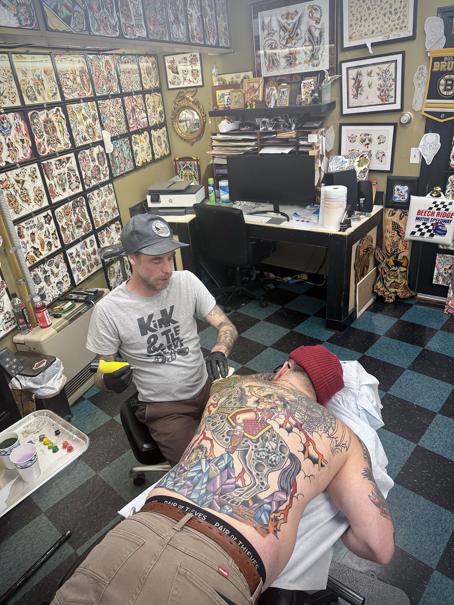 Tattoo artist tattooing a person's back in a tattoo shop filled with tattoos and artwork on the walls.