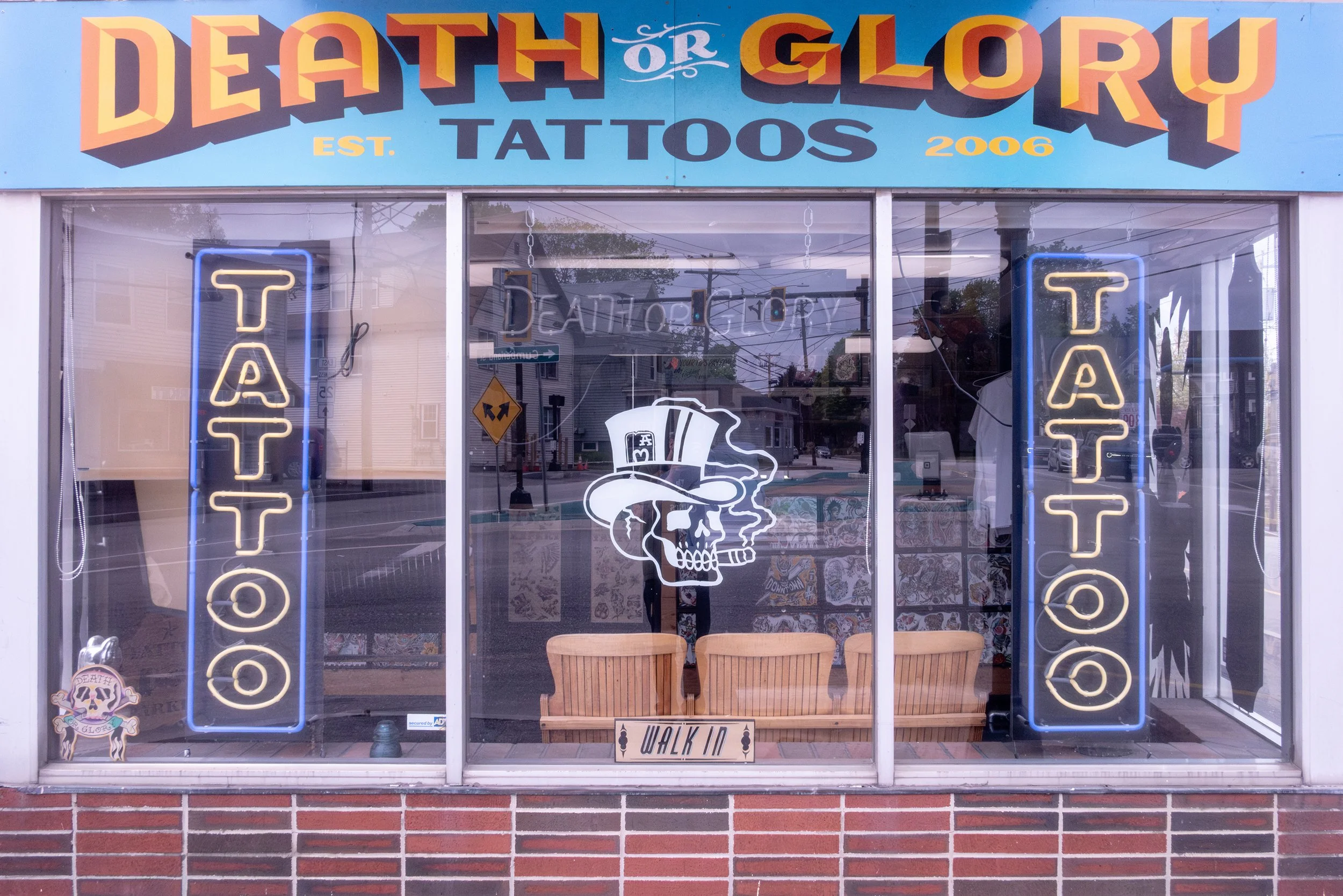 Storefront of 'Death or Glory Tattoos' with a large colorful sign above. The window displays neon signs spelling 'TATTOO' on each side, an illustration of a skull with a top hat in the center, and a small sign that says 'WALK IN'. Wooden chairs are inside, and the store was established in 2006.