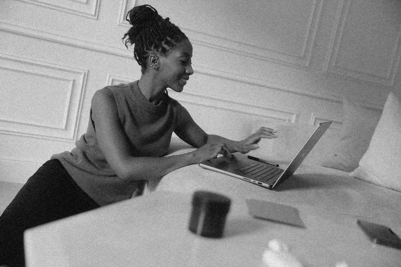 A woman with dreadlocks smiles while working on a laptop in a room with white paneled walls.