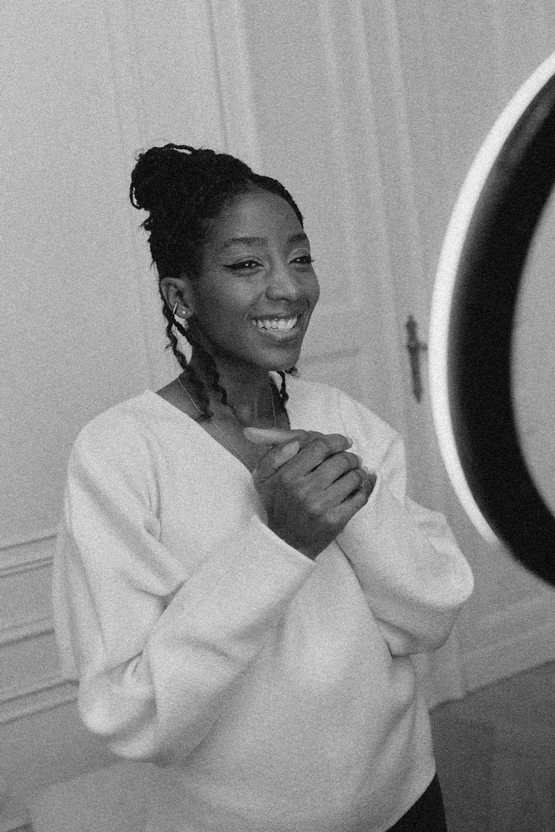 A smiling woman with braided hair, wearing a light-colored sweater, standing in front of a mirror in a room.