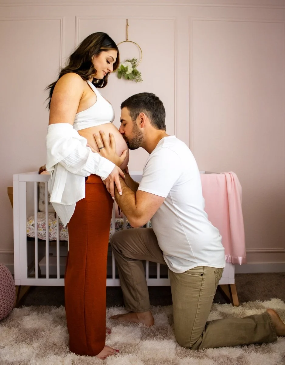 A man kisses a pregnant woman's belly while kneeling in front of her, holding her hand, in a nursery room.