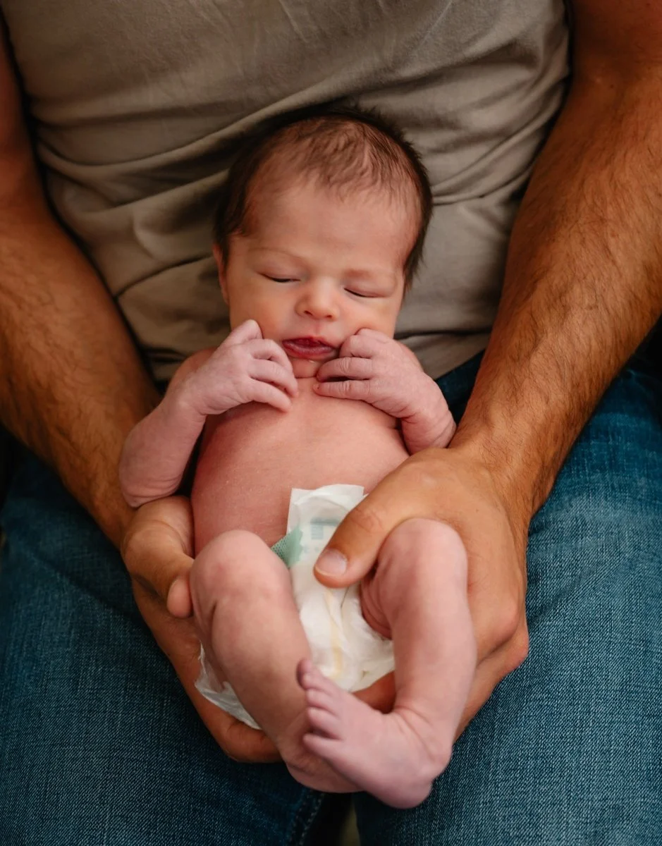 An adult sitting with a newborn baby, holding the baby securely in their lap. The baby is partially nude with a diaper, and appears to be resting or sleeping, with a calm expression and small hands near their face.