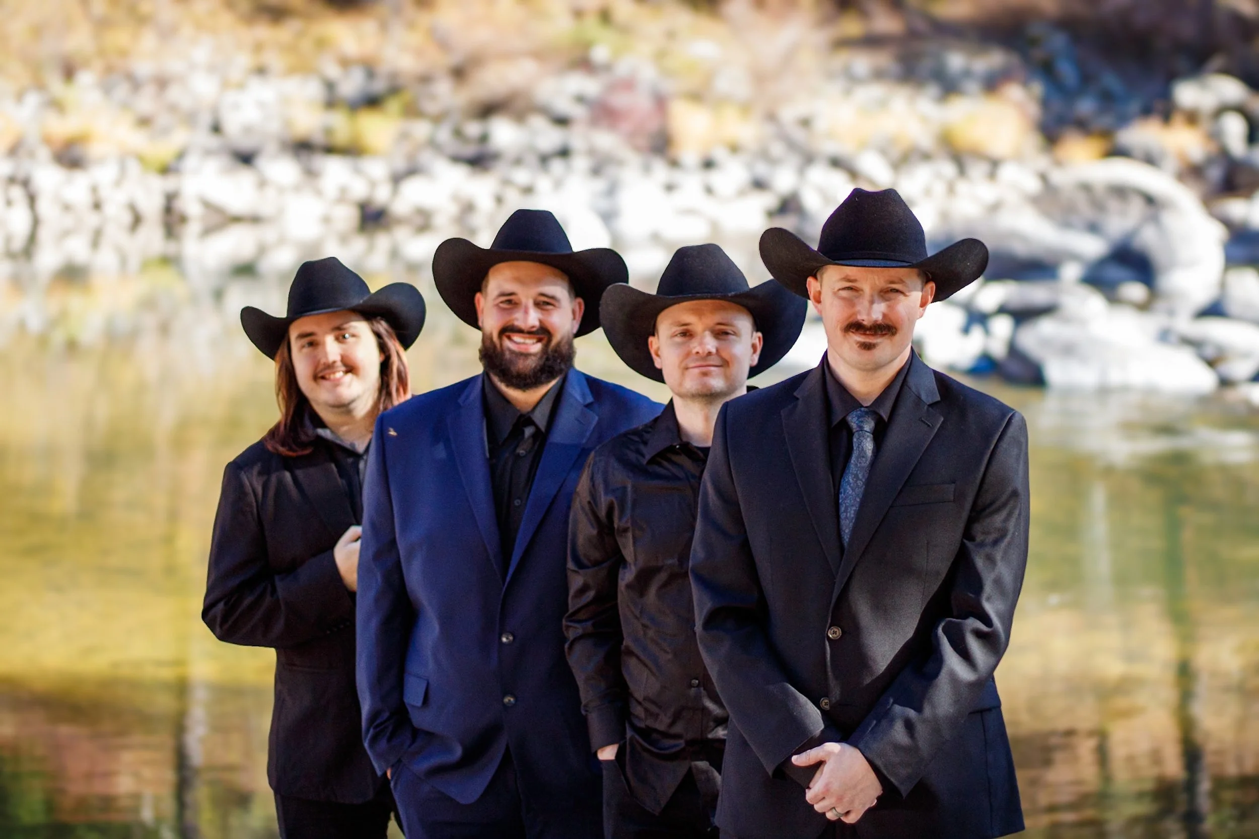 Four men and one woman wearing black suits and cowboy hats standing in front of a river and rocky shoreline.