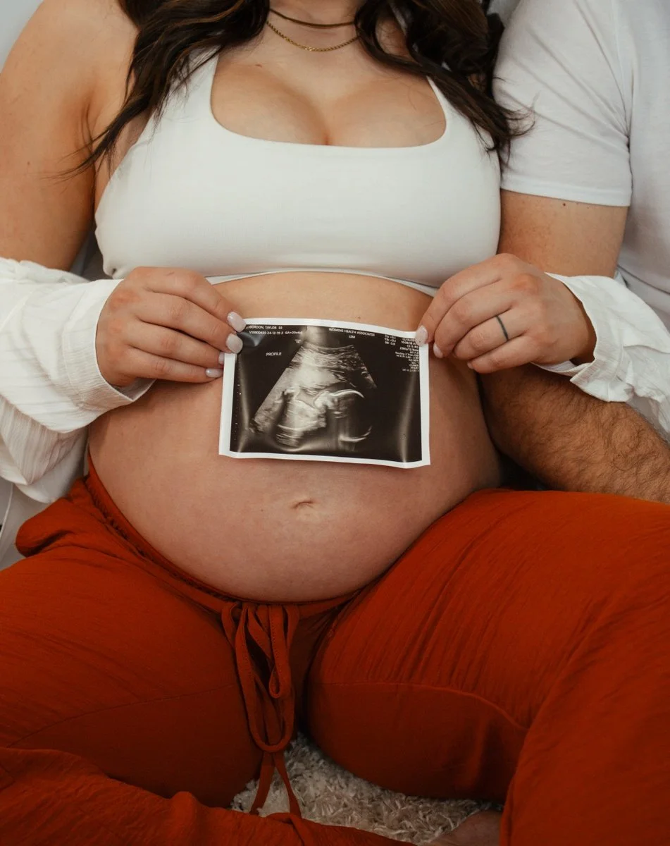 A pregnant woman holding an ultrasound image in front of her belly, sitting with her partner, who has his arm around her.