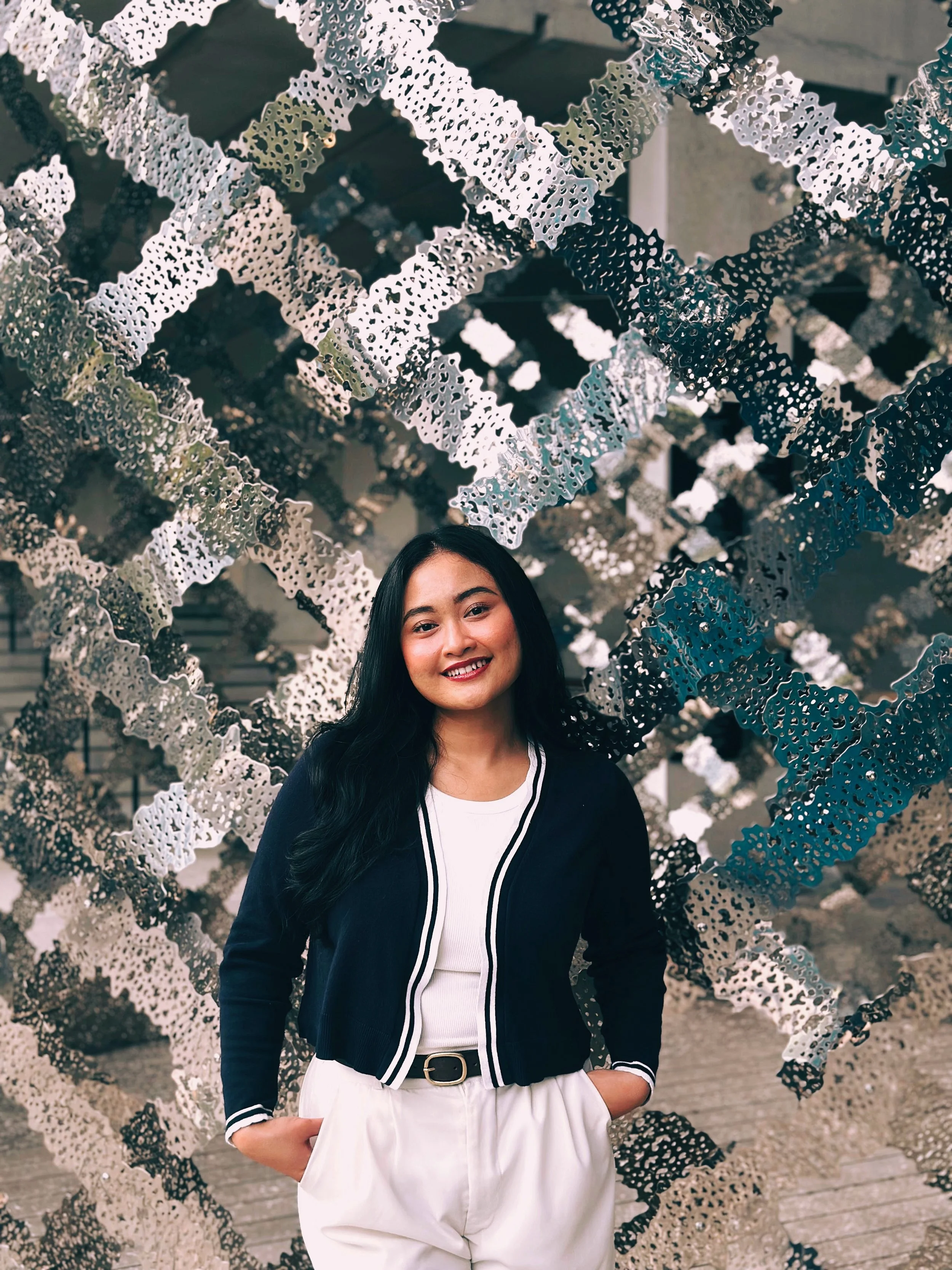 A woman with long black hair smiling and standing in front of an abstract metallic wall installation.