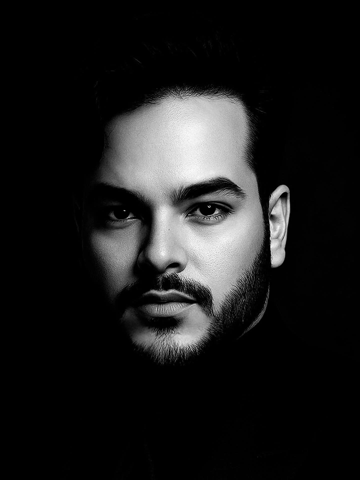 Black and white portrait of a man with dark hair, a beard, and mustache, looking directly at the camera against a black background.