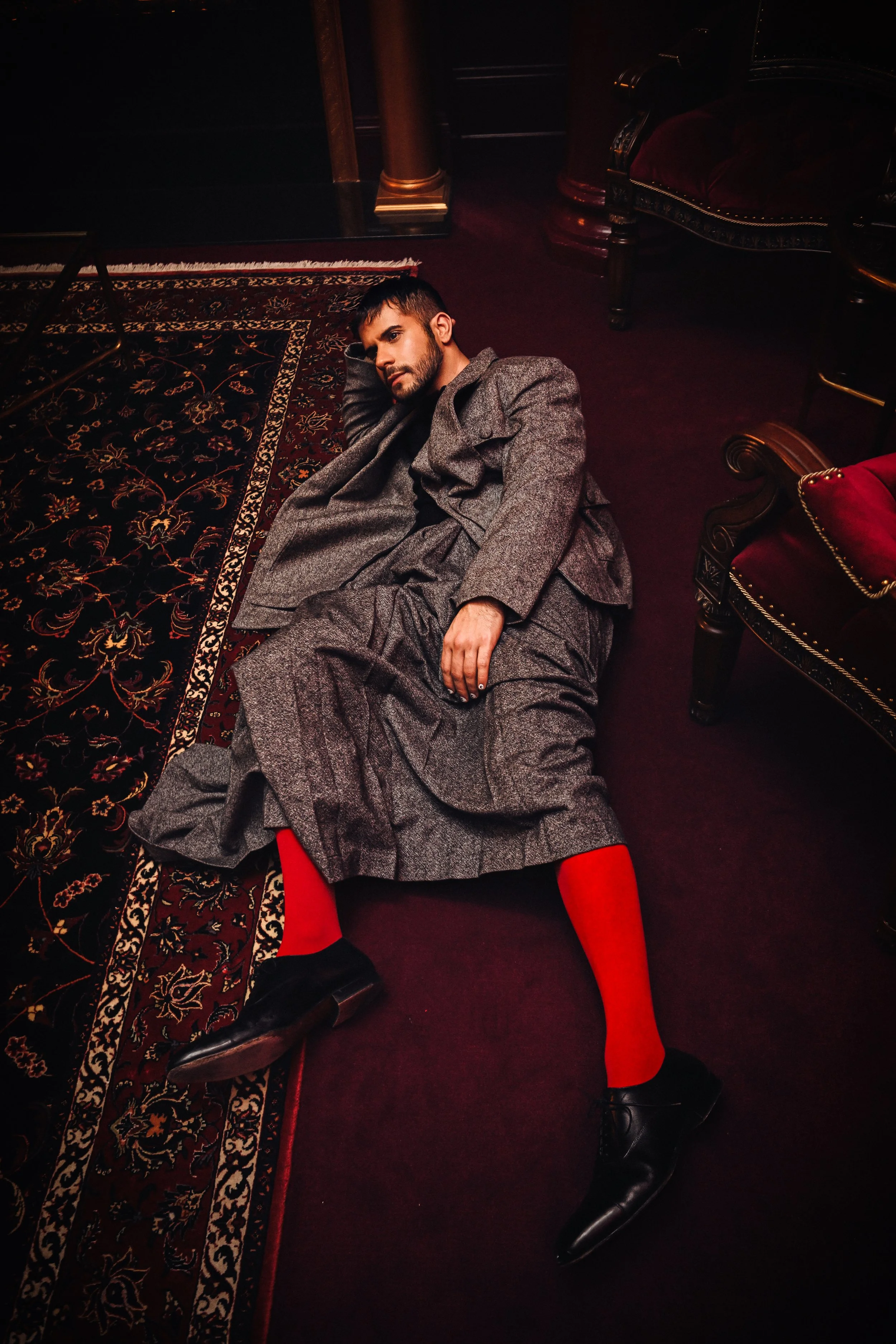 A man wearing a gray suit lying on a patterned rug on a dark red carpet in a luxurious room with vintage furniture.