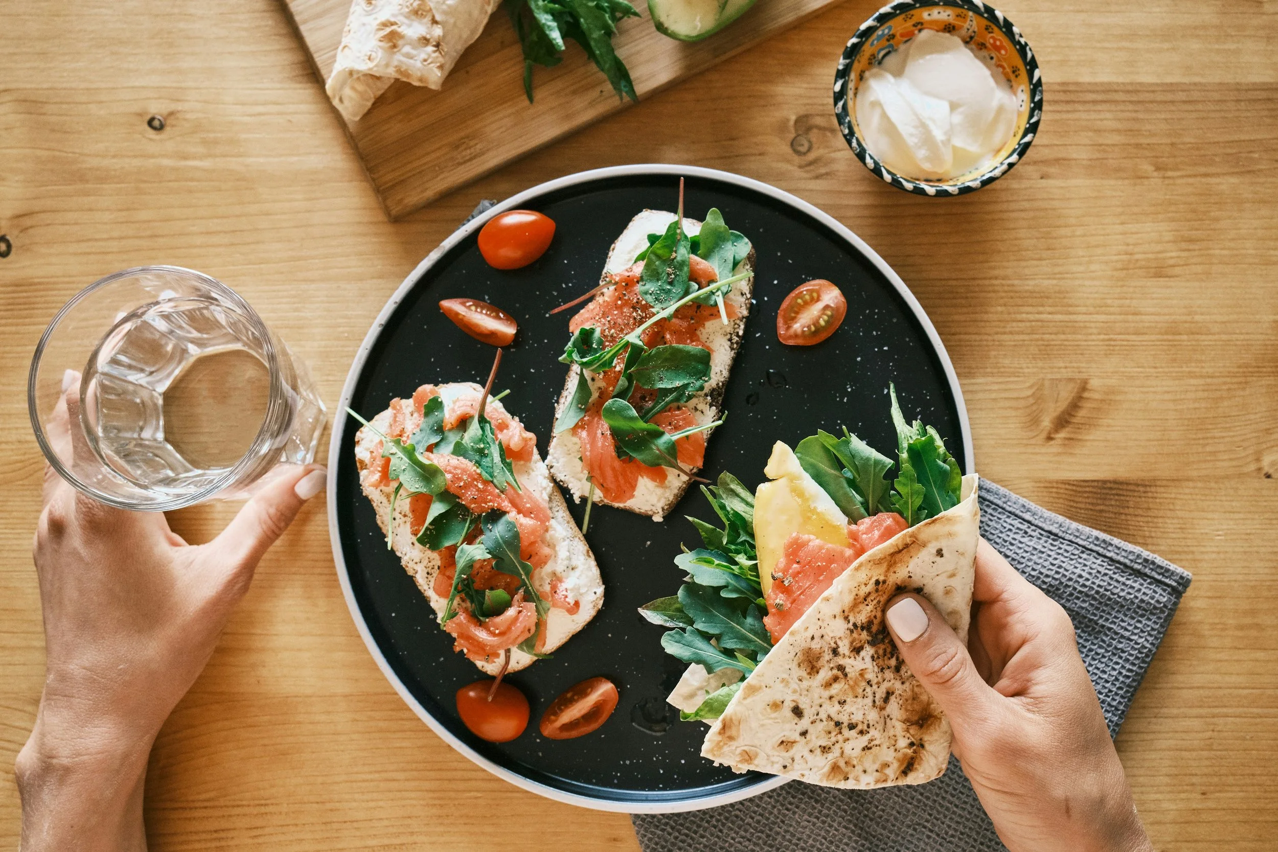 Overhead view of a black plate with two open-faced sandwiches topped with smoked salmon, arugula, and cherry tomatoes, along with a half pita bread with greens and smoked salmon. A hand holding a glass of water is visible on the left, and a hand holding the pita is on the right. There is a glass of water, cream, and a cutting board with cucumbers, herbs, and a piece of bread on a wooden table.