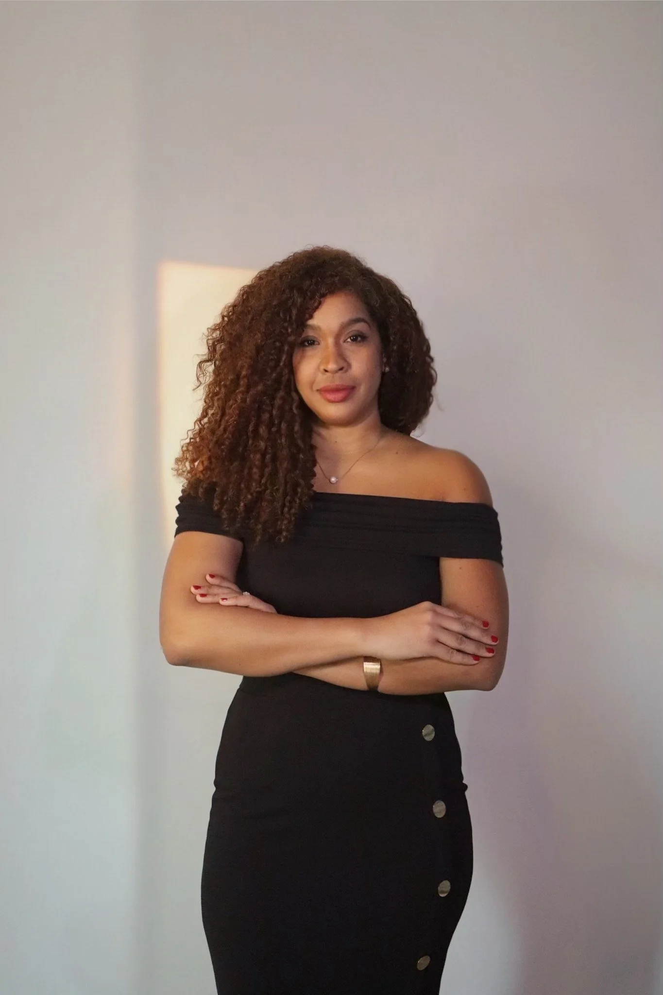 A woman with curly brown hair wearing a off-the-shoulder black dress with buttons on the side, standing with her arms crossed against a plain light-colored wall with a shadow in the background.