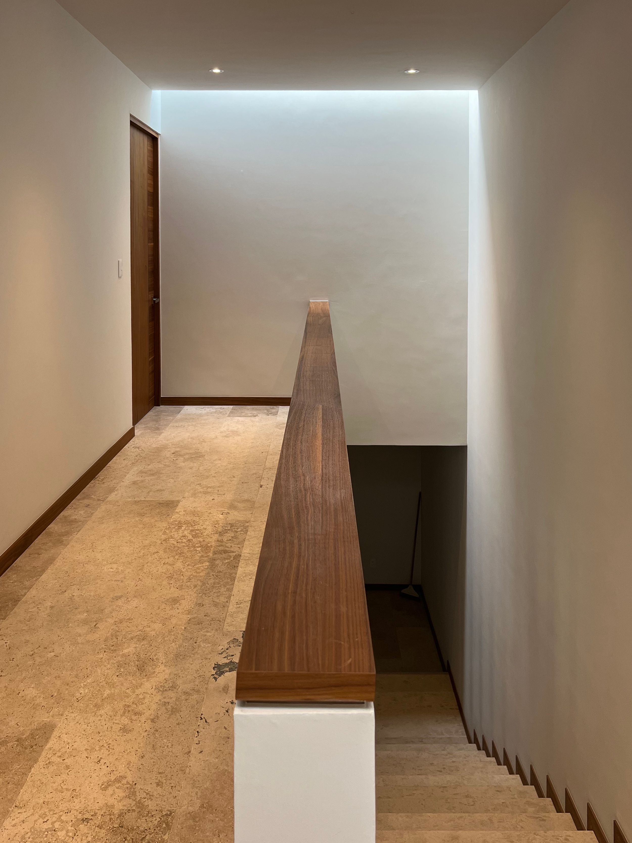 Pasillo de una casa con pared blanca, barandal de madera y escaleras hacia abajo.
