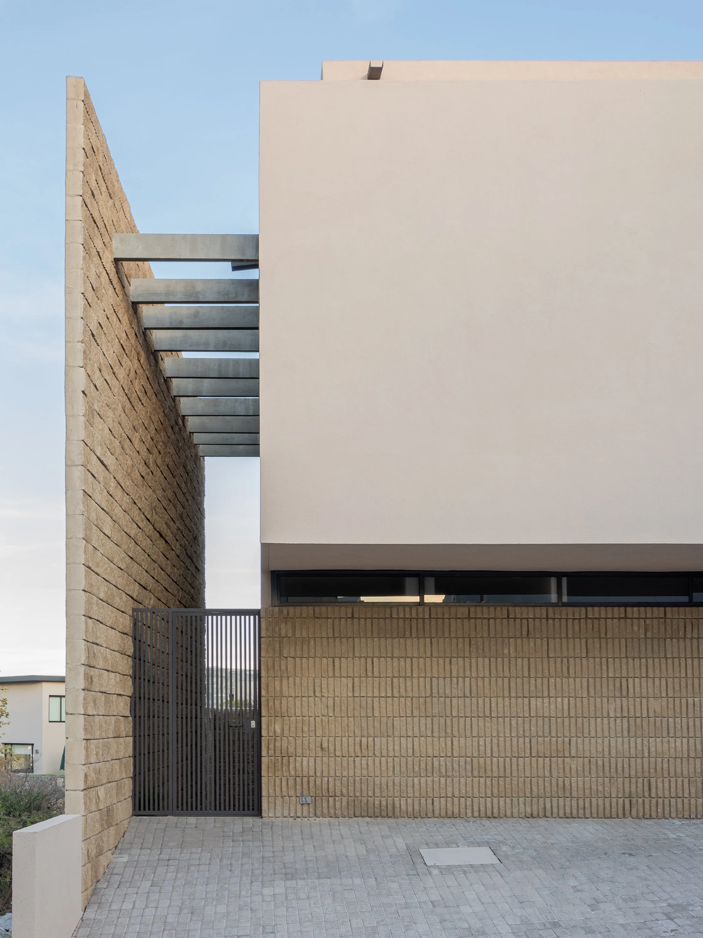 Vista frontal de una casa moderna con paredes de ladrillo y una pared blanca, con una puerta metálica negra en la parte inferior izquierda y ventanas estrechas en la parte superior, en un día despejado.
